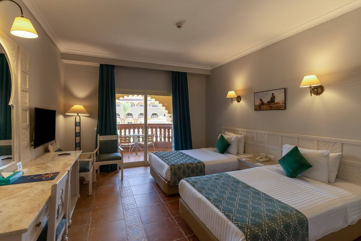 Habitación de hotel con dos camas, un escritorio con silla, televisor y balcón con vistas a un edificio.