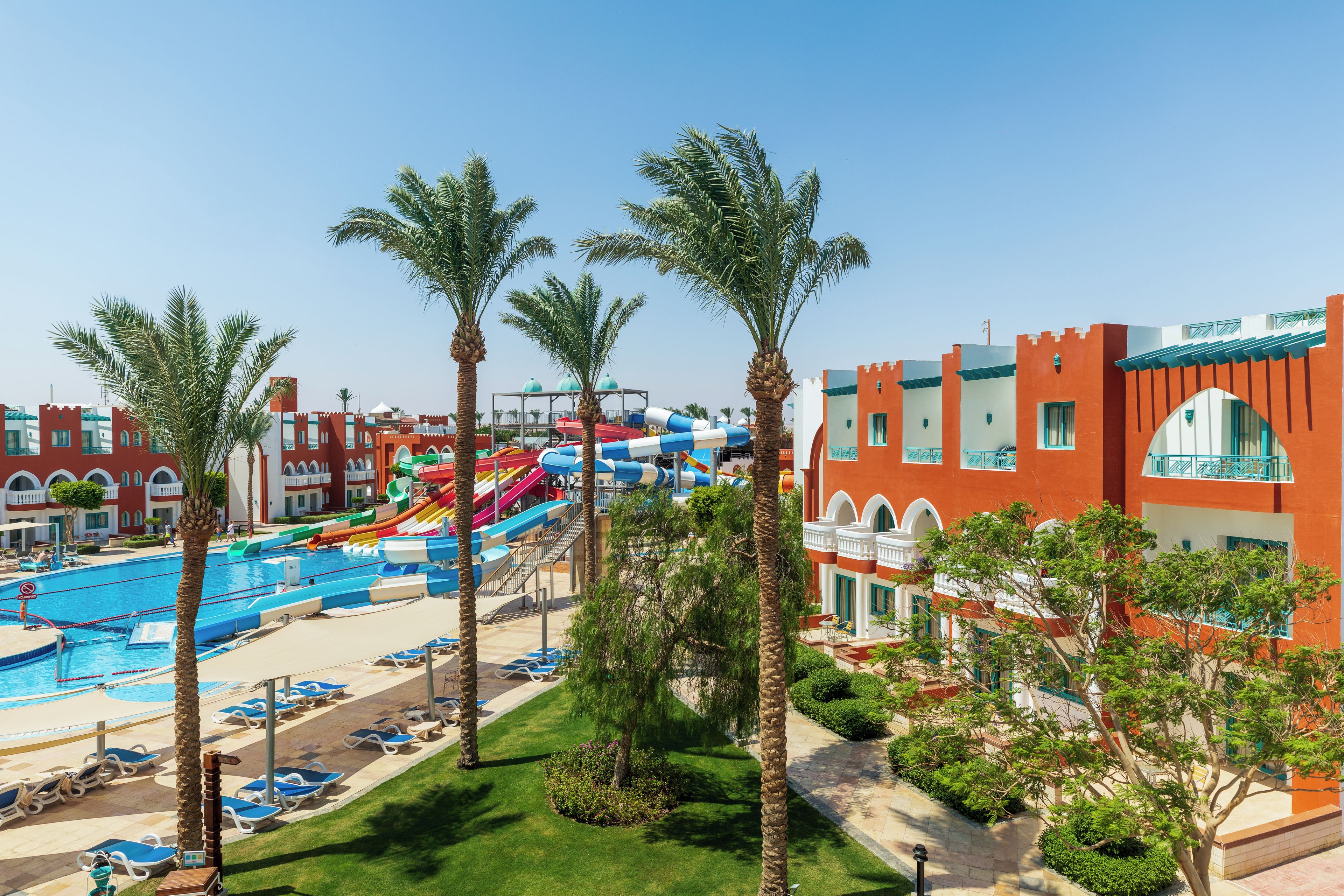 A resort with a swimming pool, water slides, palm trees, and a building with a red facade.