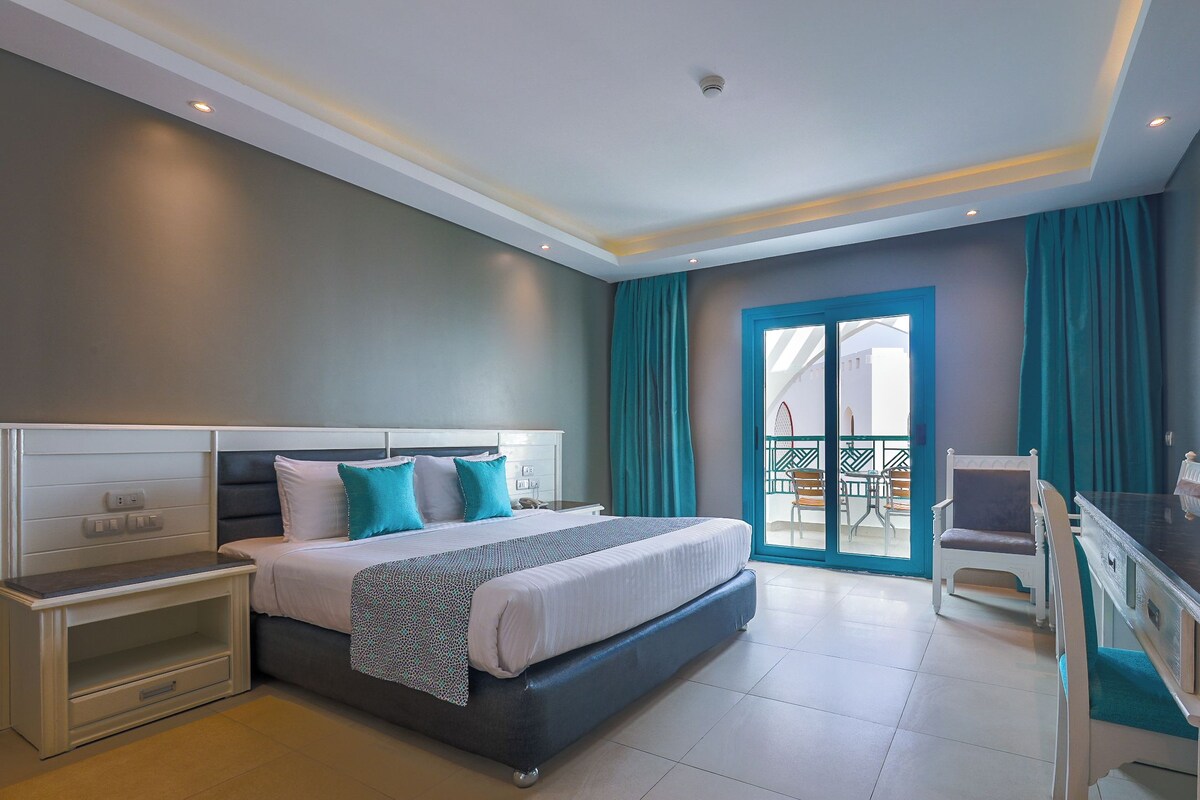 A modern hotel room with a large bed, a desk, and a balcony with a view.