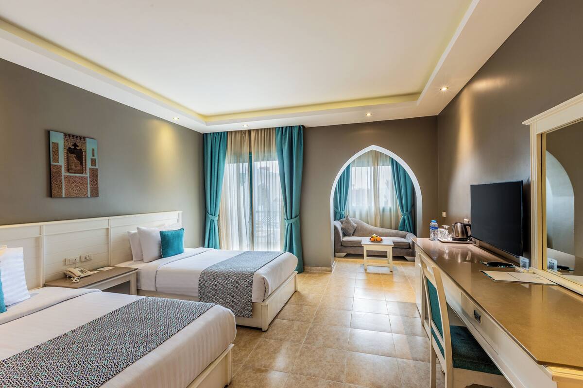 Habitación de hotel con dos camas, un escritorio, un televisor y vistas a la ciudad a través de amplios ventanales.