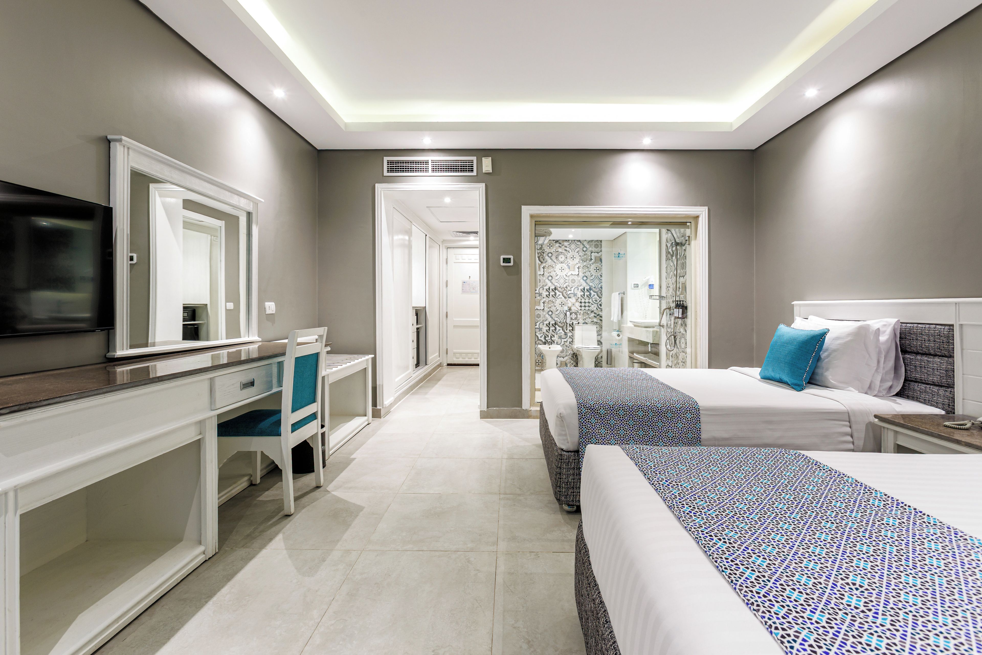 Ein modernes Hotelzimmer mit Flachbildfernseher, Schreibtisch mit Stuhl, Bett mit gemusterten Betttextilien und einem durchgehenden Türspalt sichtbaren Badezimmer.