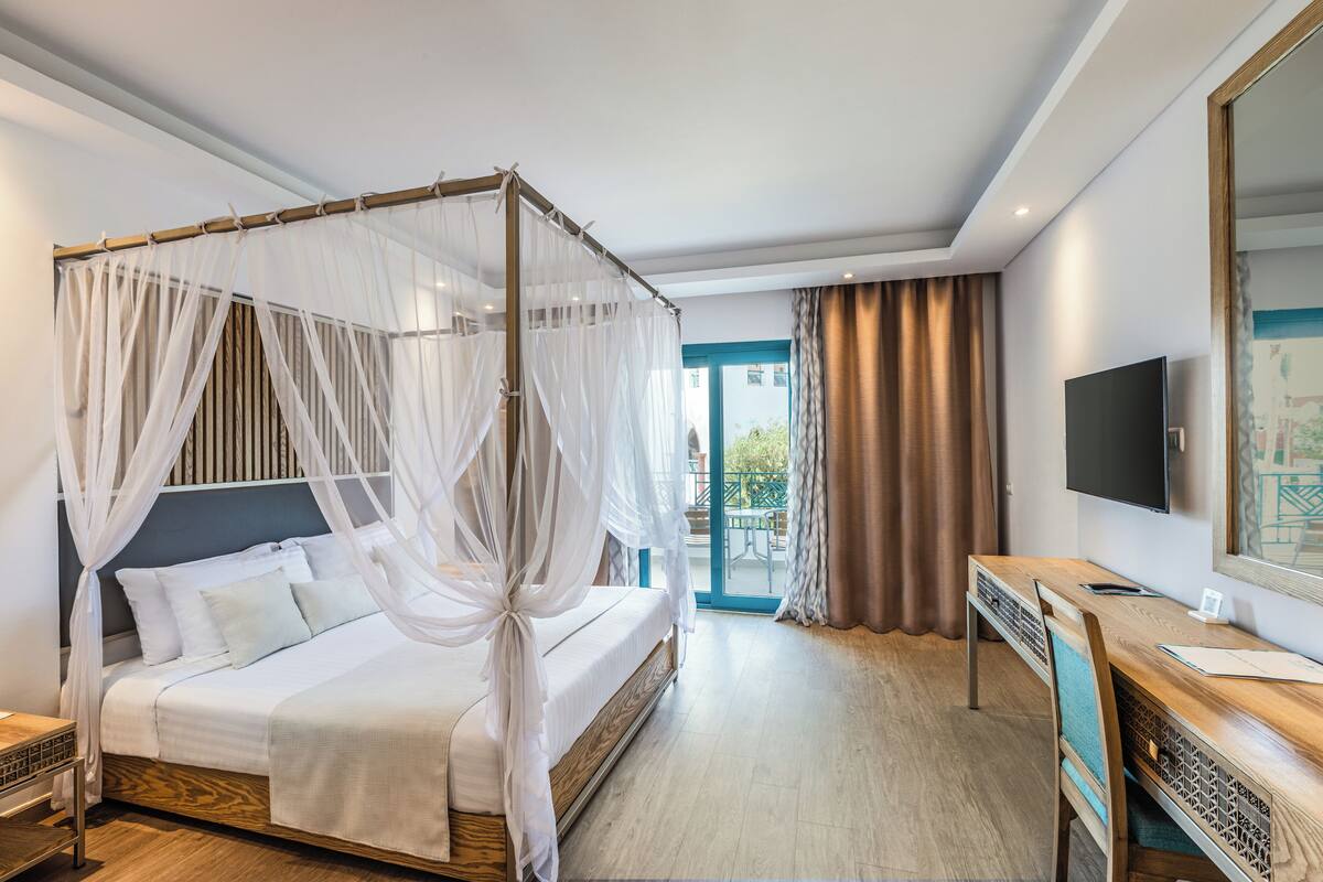 Una habitación de hotel con una cama con dosel, un escritorio con silla, un televisor de pantalla plana y un ventanal con cortinas.