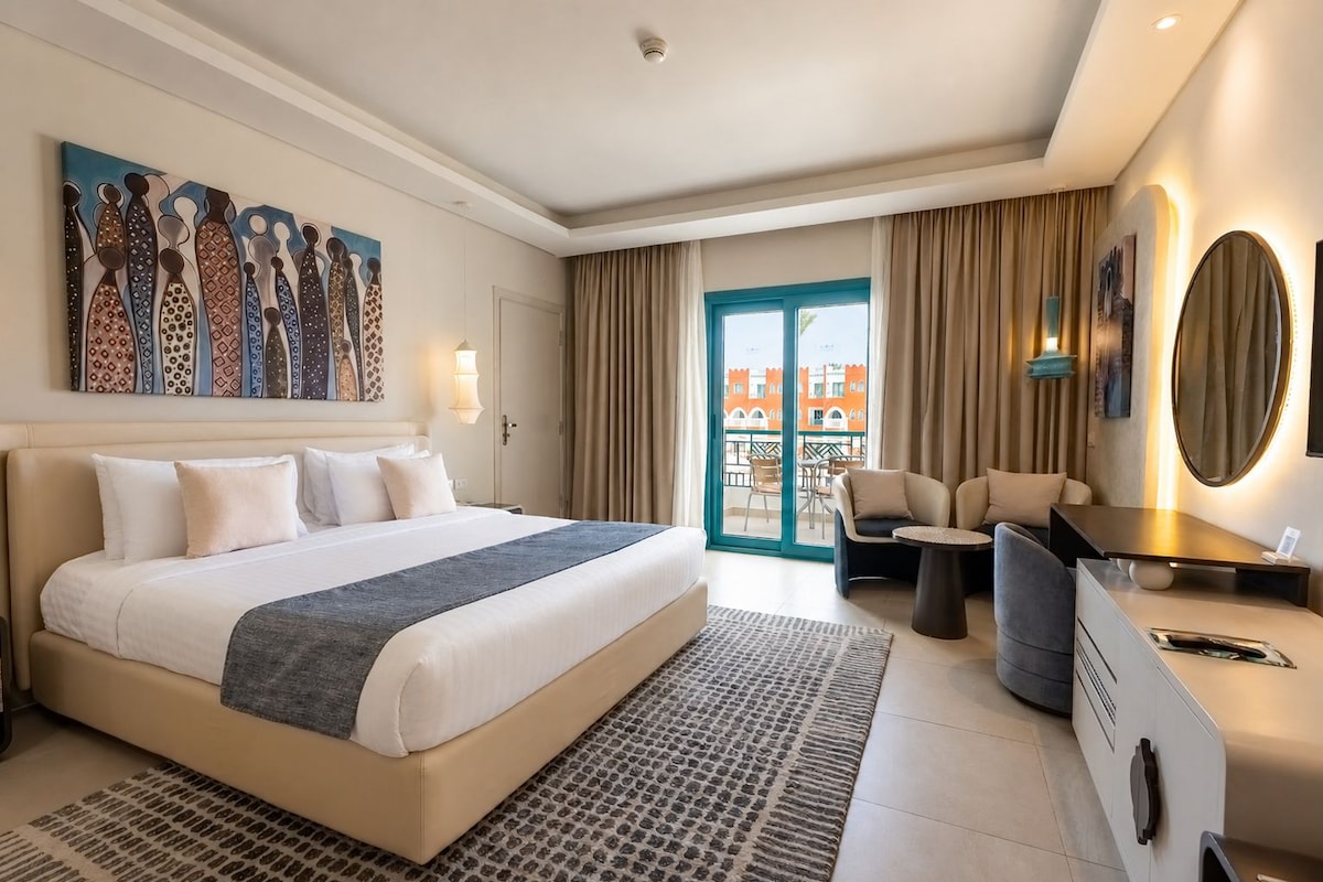 A hotel room with a large bed, a seating area with a sofa and chairs, a desk, a large mirror, and a view of buildings through the window.