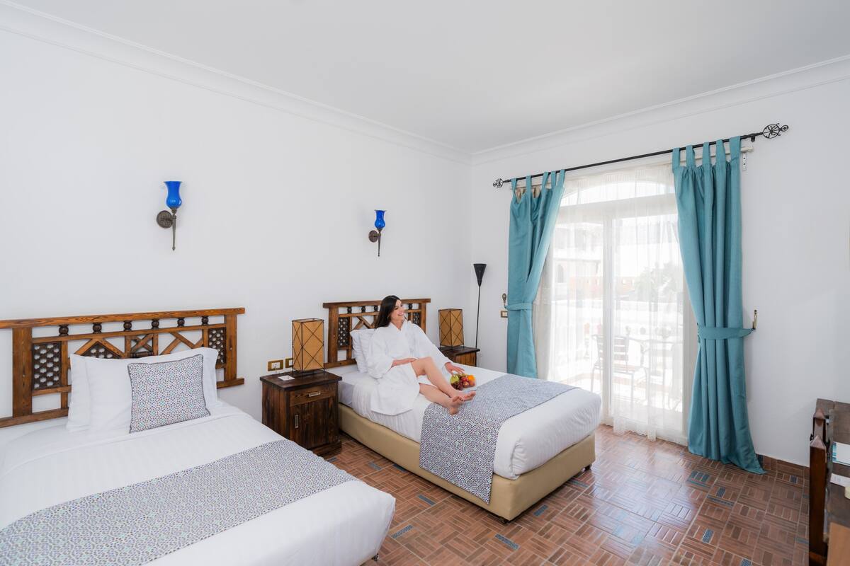 Habitación de hotel con dos camas, cabecera de madera, un ventanal grande con cortinas azules y vistas al exterior.