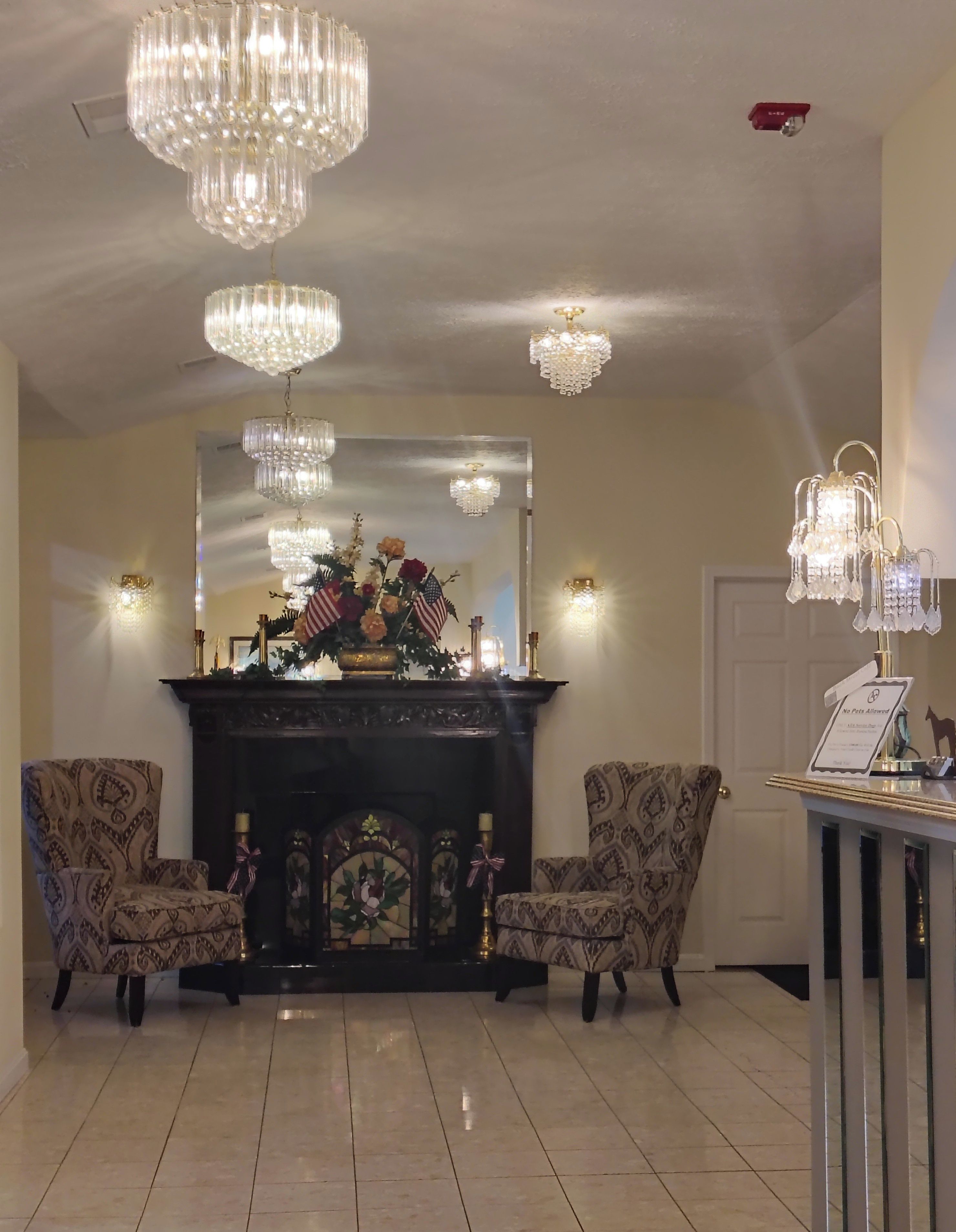 A room with a fireplace, two patterned armchairs, a mirror, and multiple chandeliers.