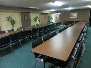 Un salón de conferencias con mesas largas y sillas, alfombra verde y plantas en macetas.