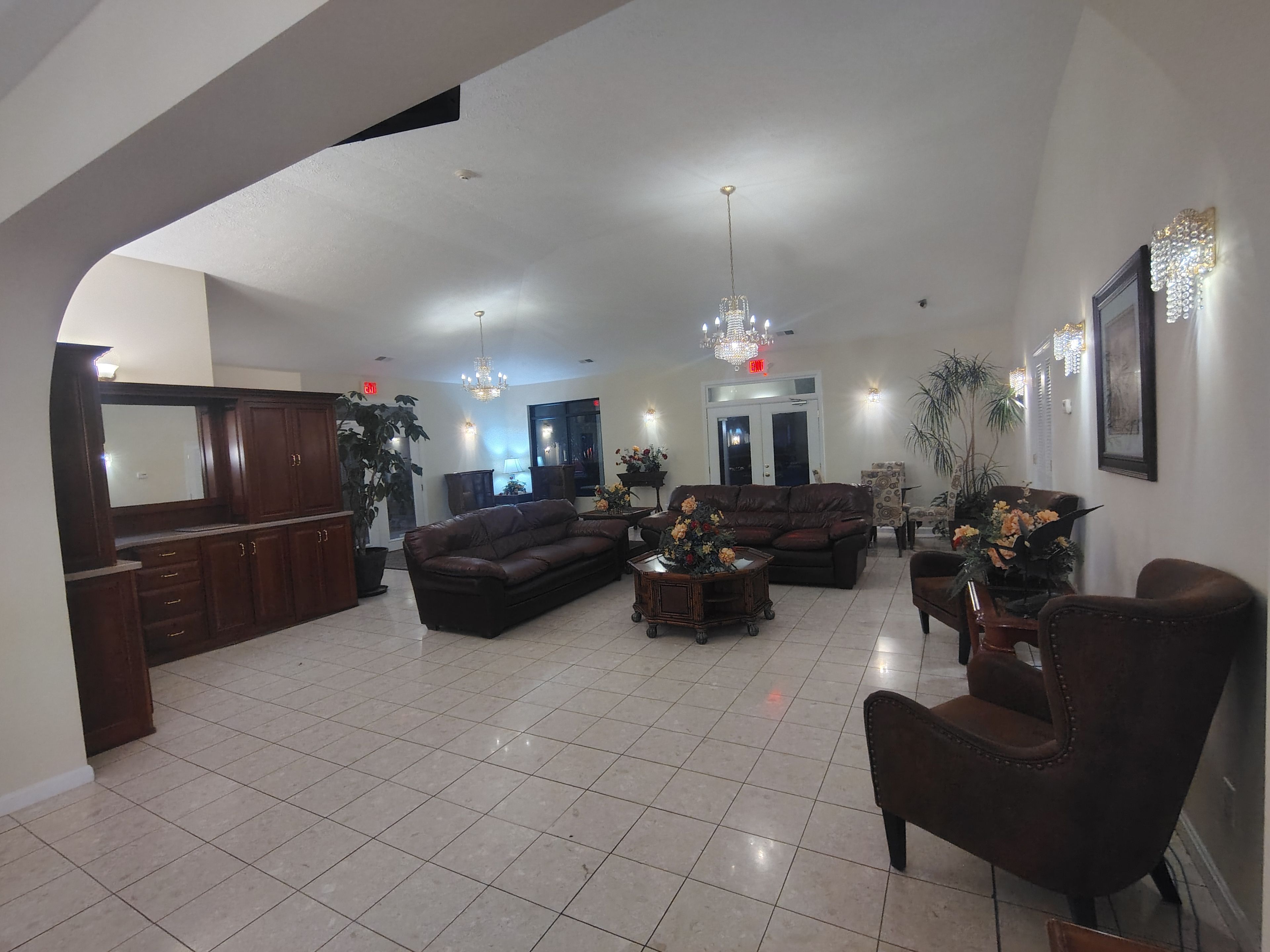 A spacious living area with a large sofa, armchairs, and a coffee table. There is a chandelier hanging from the ceiling, and the room has a tiled floor.