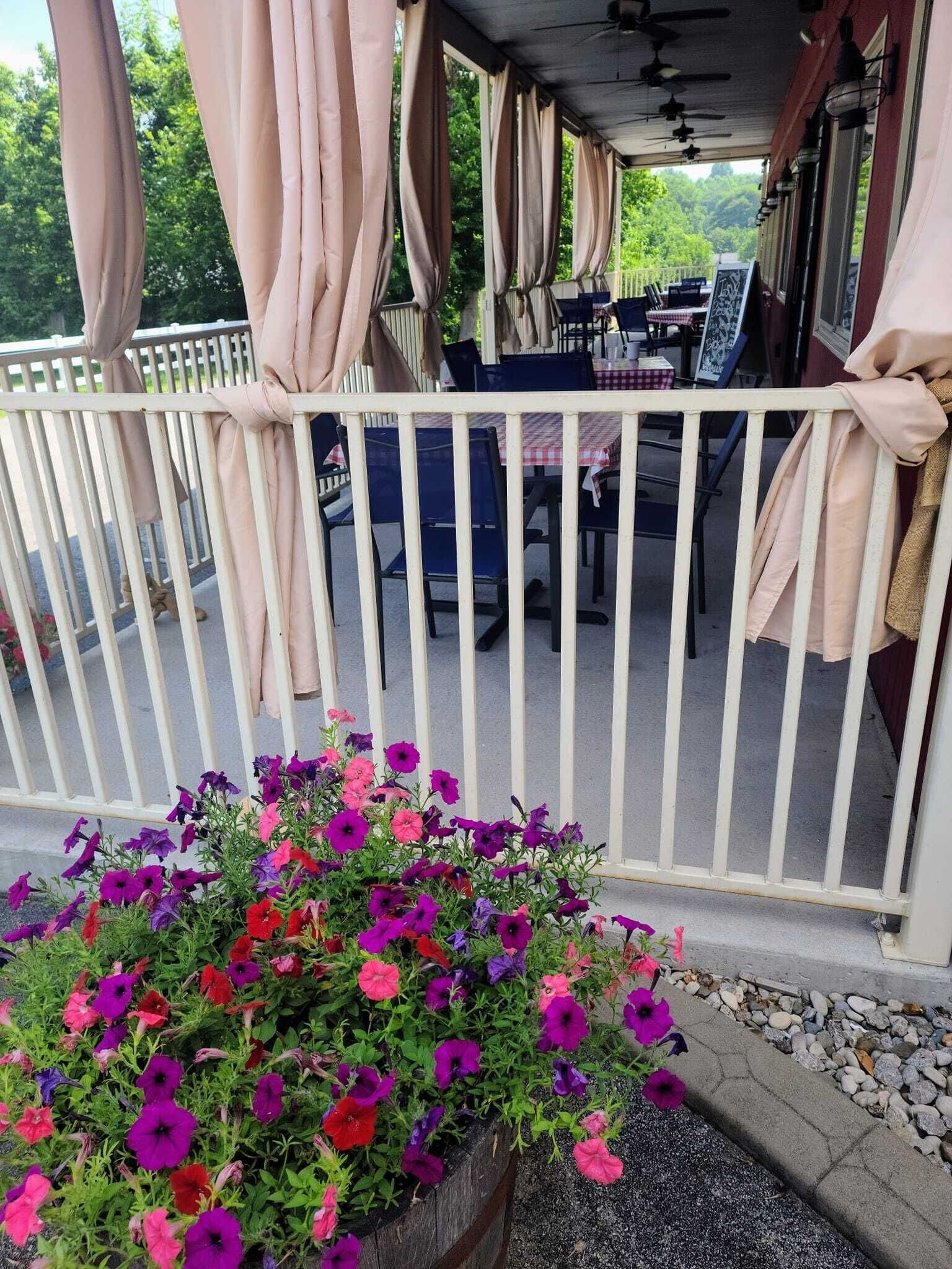 Un espacio al aire libre con techo, mesas y sillas, una cerca blanca y una jardinera con flores de colores.