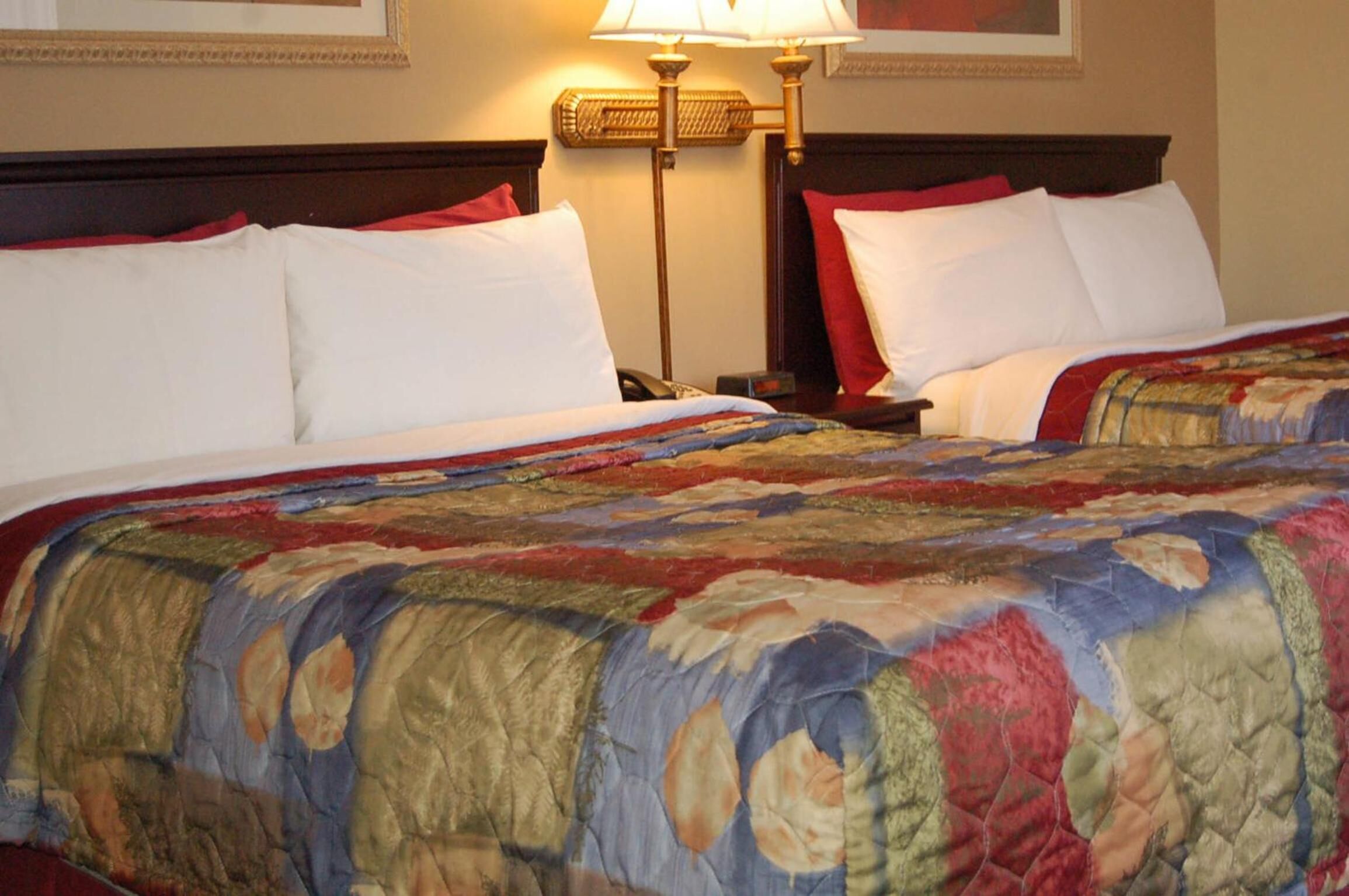 A hotel room with two beds, each with white bedding and red pillows. There is a large, colorful quilt on the beds. A lamp is on the nightstand beside each bed.