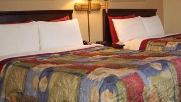 A hotel room with two beds, each with white bedding and red pillows. There is a large, colorful quilt on the beds. A lamp is on the nightstand beside each bed.