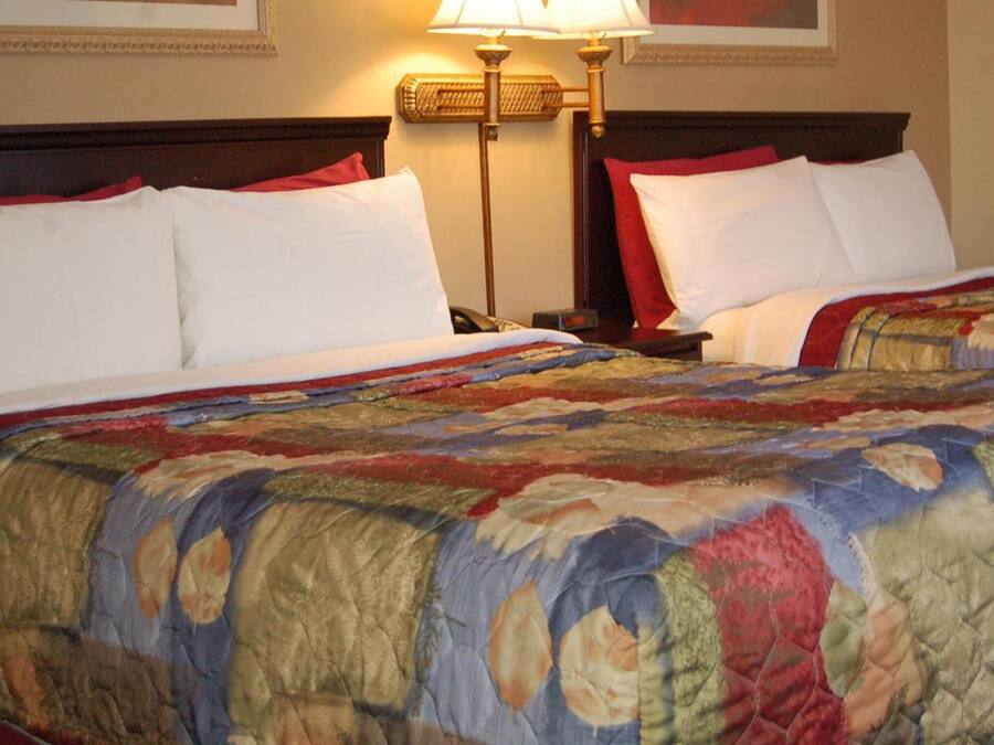A hotel room with two beds, each with white bedding and red pillows. There is a large, colorful quilt on the beds. A lamp is on the nightstand beside each bed.