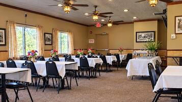 A conference room with multiple tables and chairs, large windows with curtains, ceiling fans, and framed pictures on the walls.
