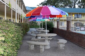 Outdoor tables and benches with a colorful umbrella, a brick wall, and a building with balconies in the background.