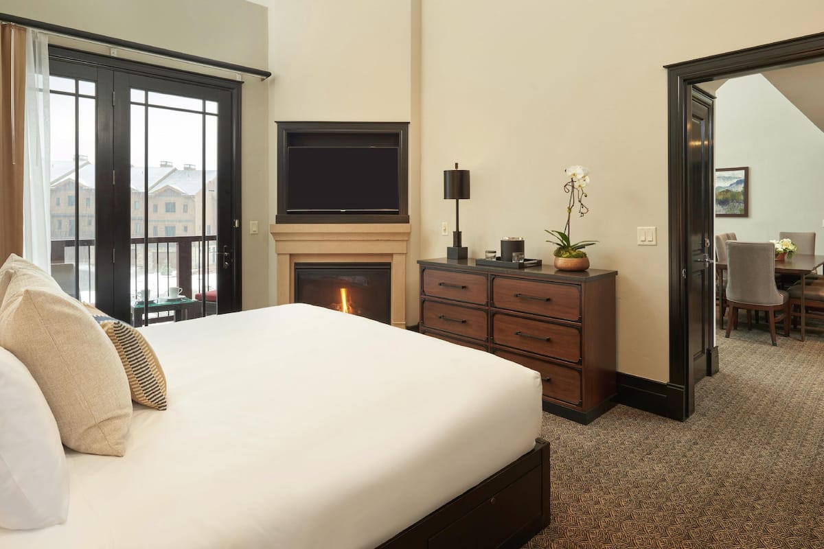 A hotel room with a large bed, a fireplace, a TV, and a view of buildings through the window.
