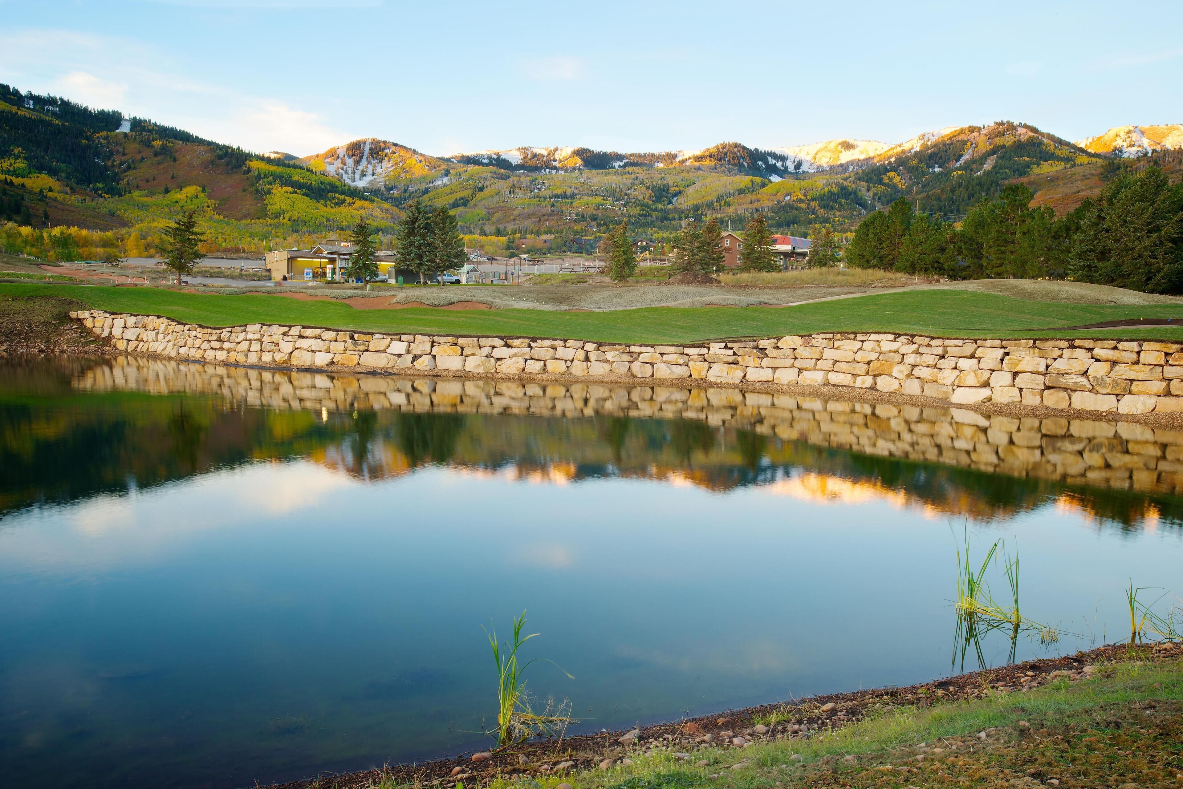 Un muro de piedra rodea un lago tranquilo que refleja el paisaje circundante, incluyendo montañas y algunos edificios al fondo.