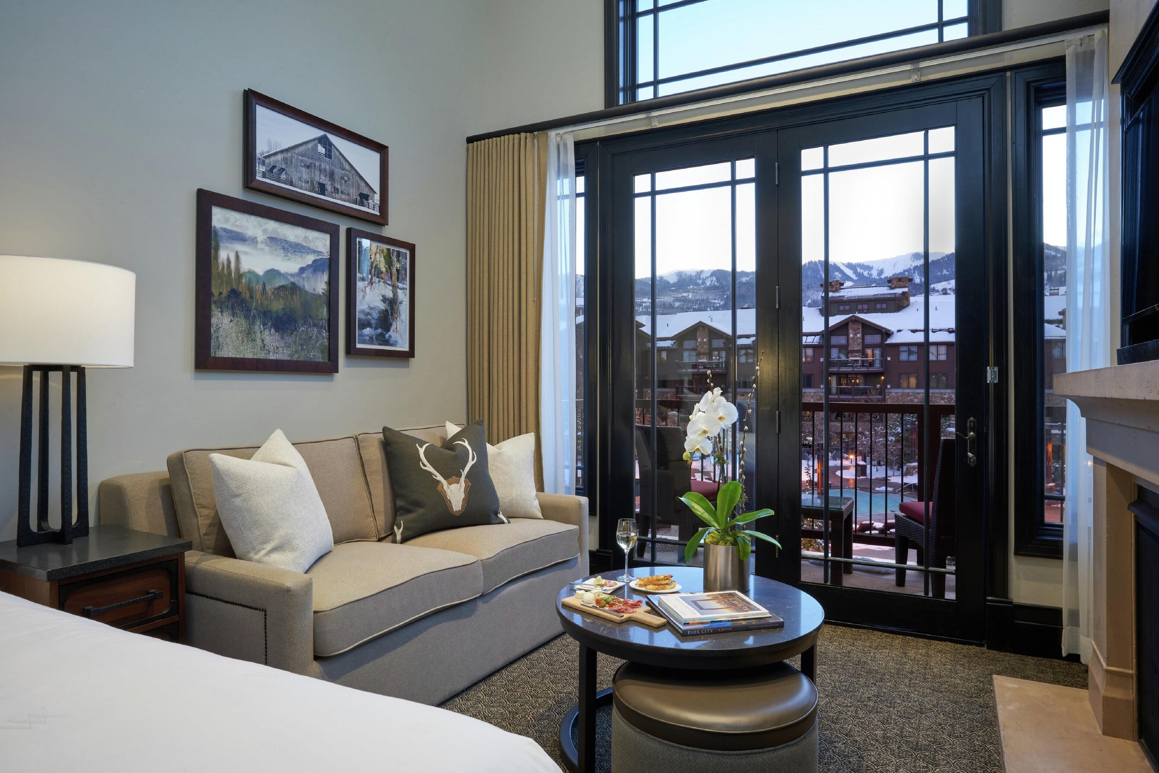 A hotel room with a sofa, a dining table with a vase of flowers, a lamp, and a view of a mountain resort through the sliding glass door.