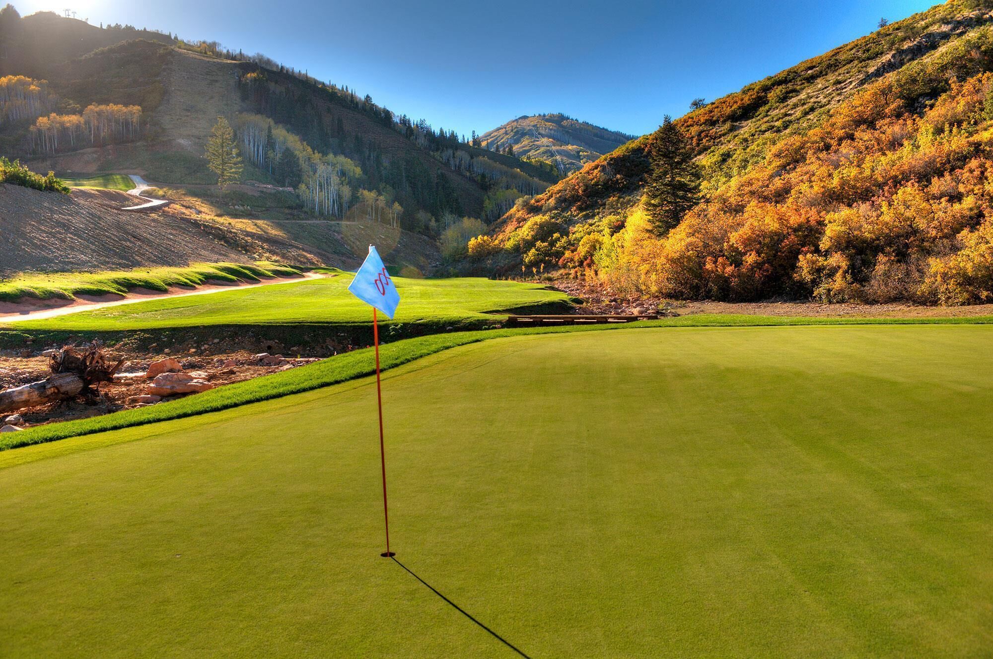 Un campo de golf con una bandera en el green, rodeado de árboles y colinas de colores otoñales.