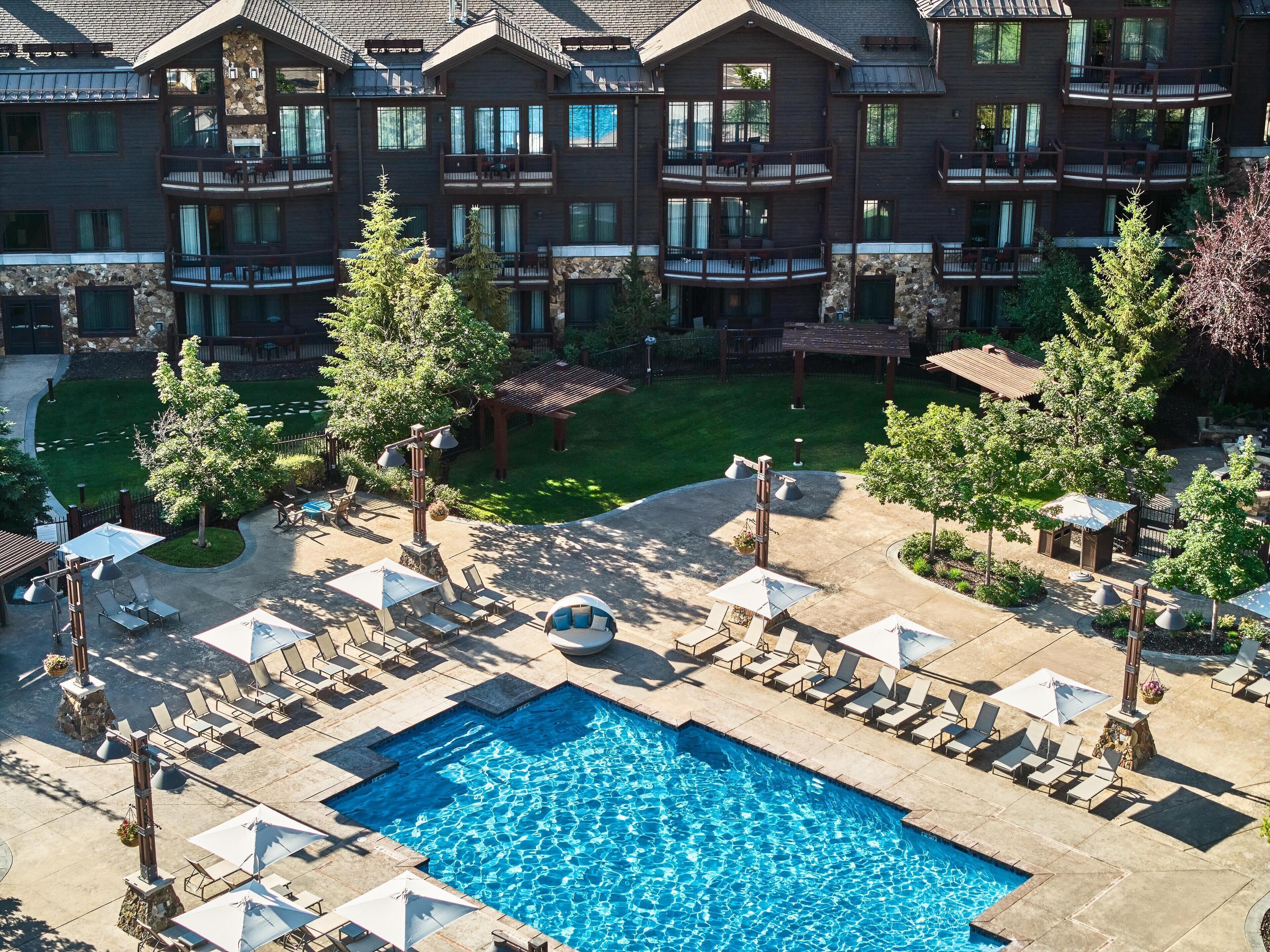 A hotel with a swimming pool surrounded by lounge chairs and umbrellas, a well-maintained lawn, and a multi-story building with balconies.