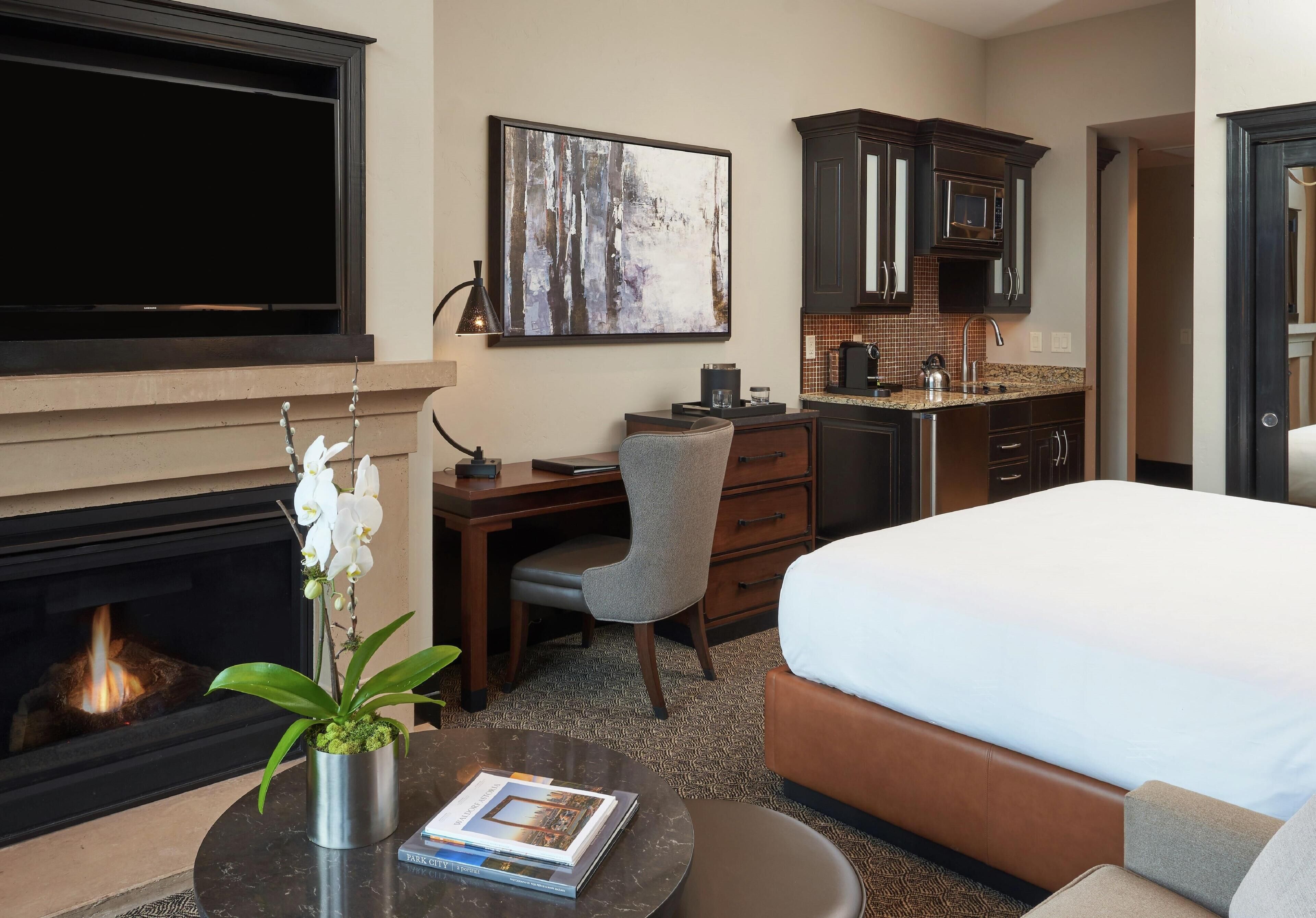 Een hotelkamer met een bed, een bureau met stoel, een open haard, een televisie en een keukenhoek.