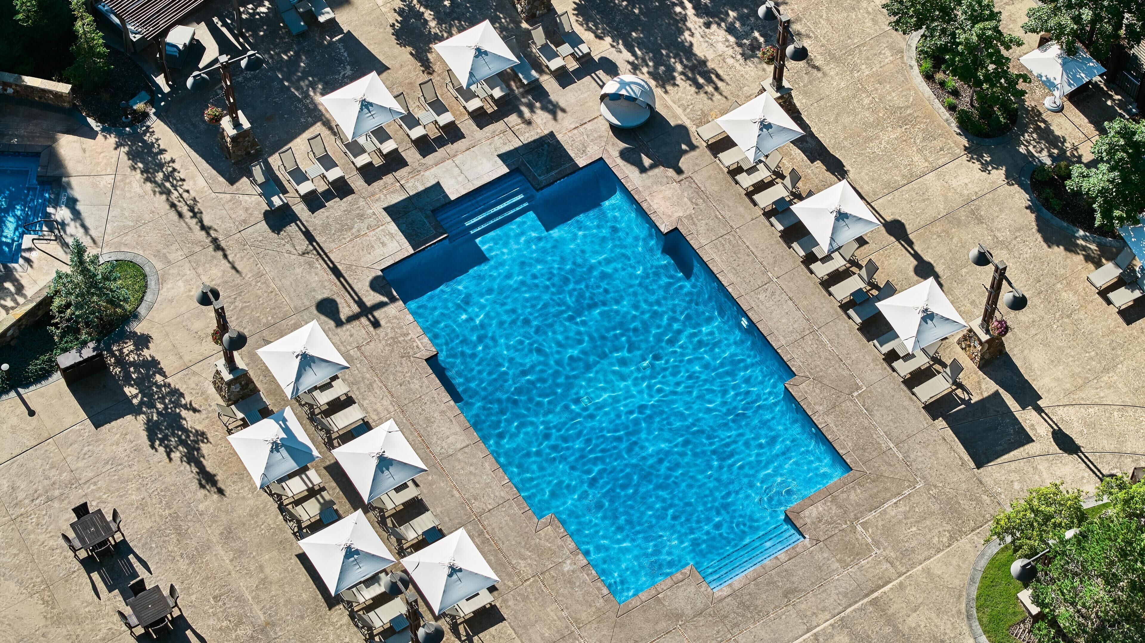 An aerial view of a swimming pool area with multiple lounge chairs and umbrellas, surrounded by a landscaped garden.