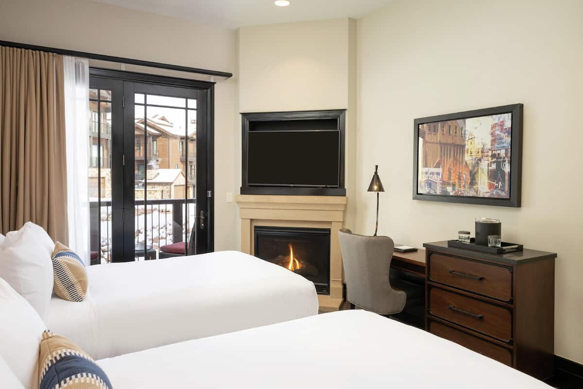 A hotel room with two beds, a fireplace, a TV, a desk with drawers, and a painting on the wall.