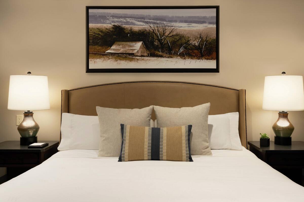 A neatly made bed with two pillows, a framed landscape painting above it, and two bedside lamps on either side.