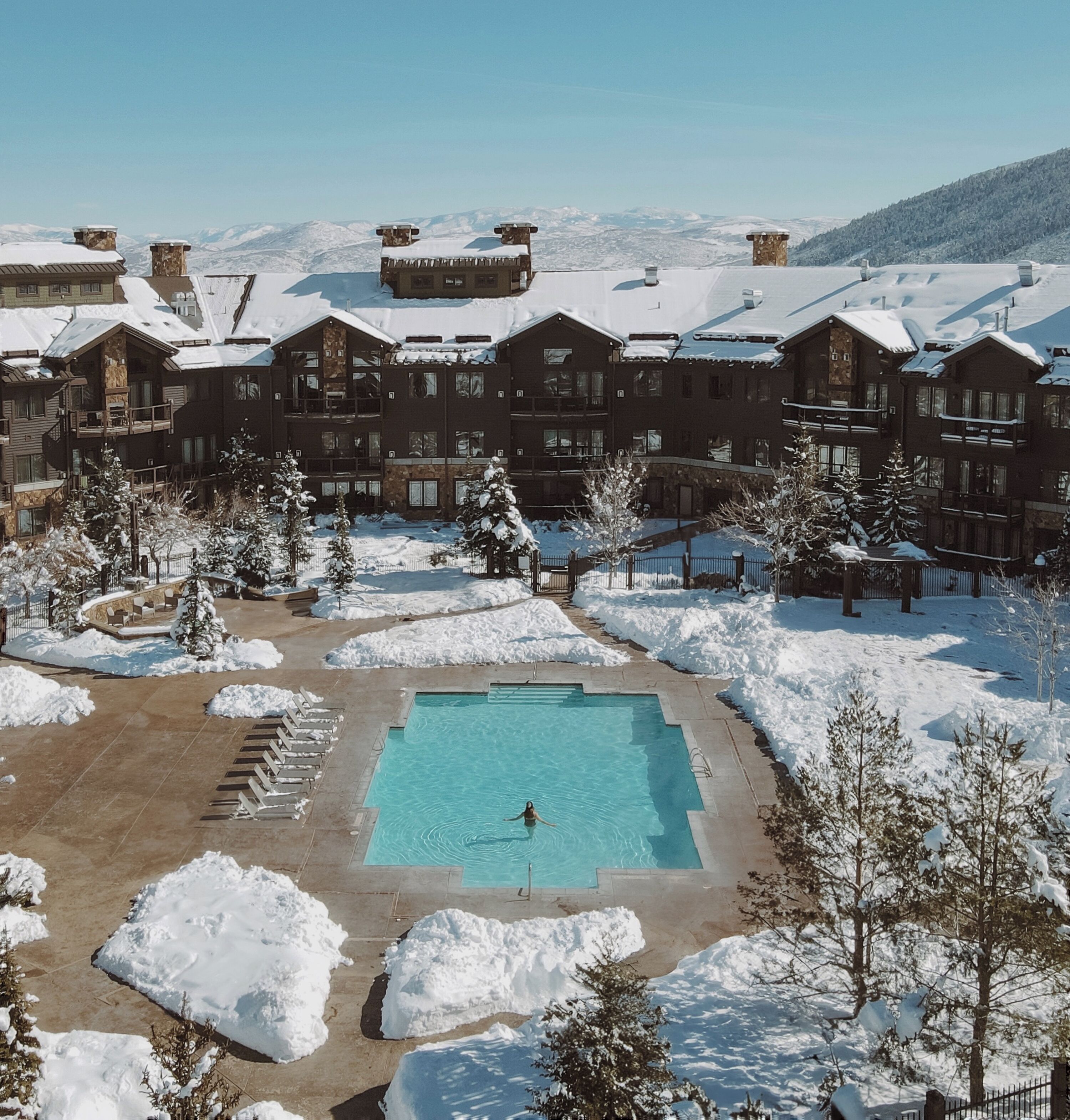 A resort with a swimming pool surrounded by snow, multiple buildings with balconies, and a mountainous backdrop.