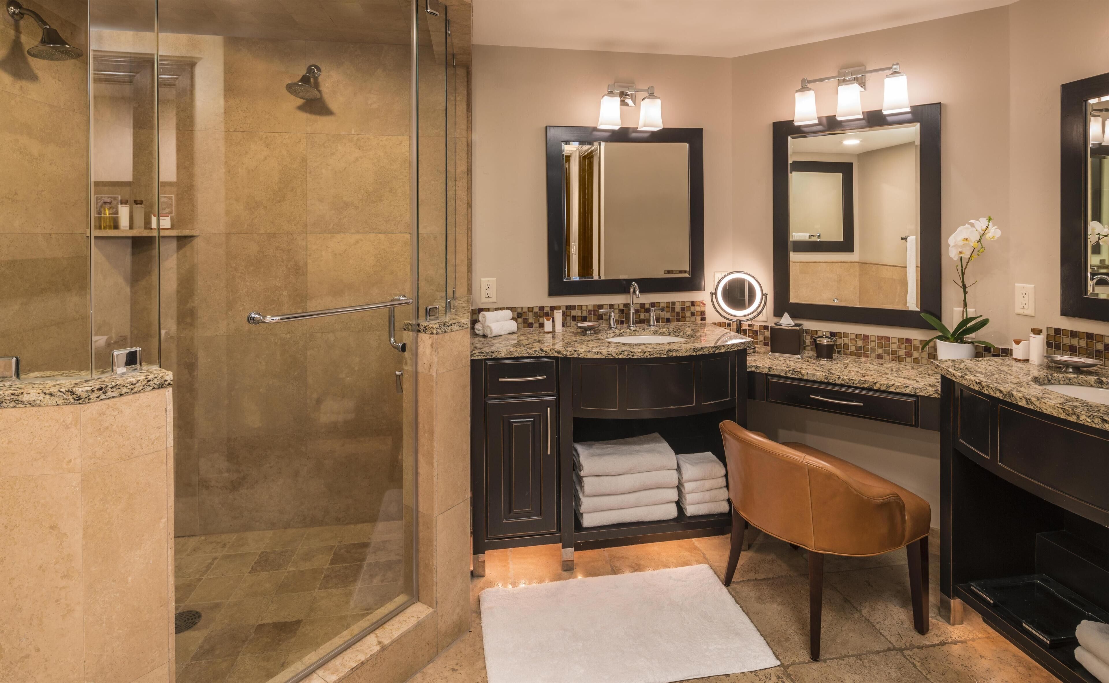 A modern bathroom with a glass shower enclosure, dual sinks, dark cabinetry, and a bench.