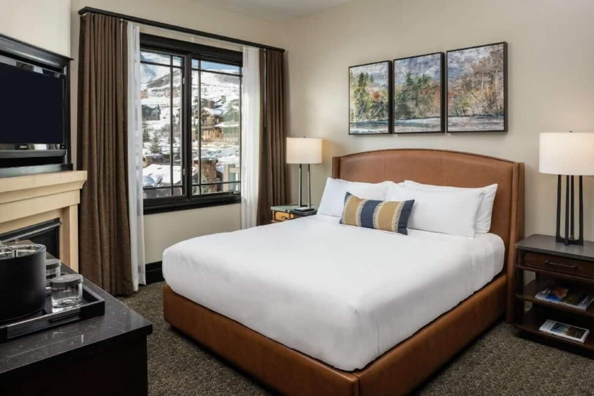 A hotel room with a large bed, two bedside lamps, a flat-screen TV, a fireplace, and a window with a view of a snowy landscape.