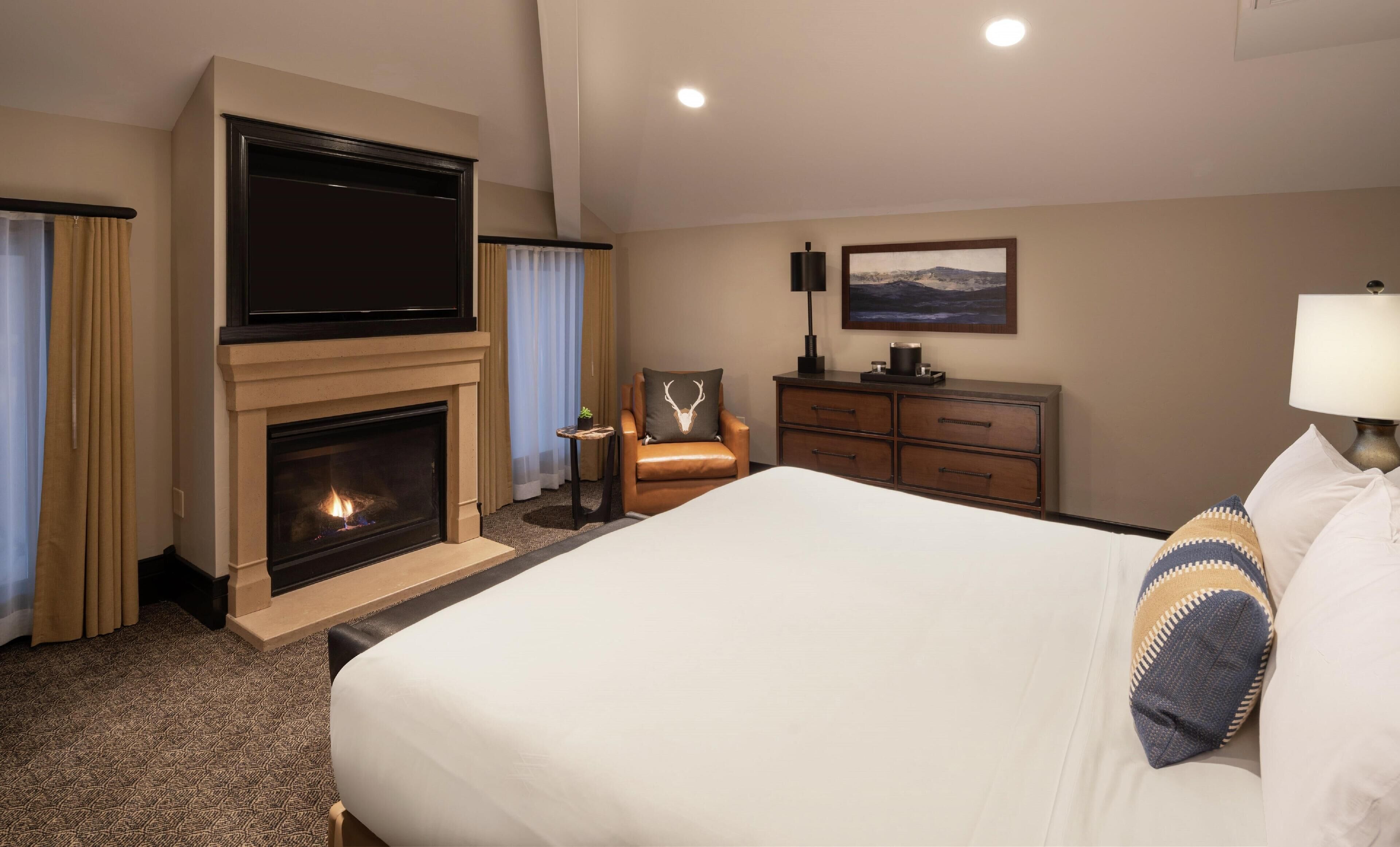 Habitación de hotel con chimenea, cama, sillón, cómoda y un cuadro en la pared.