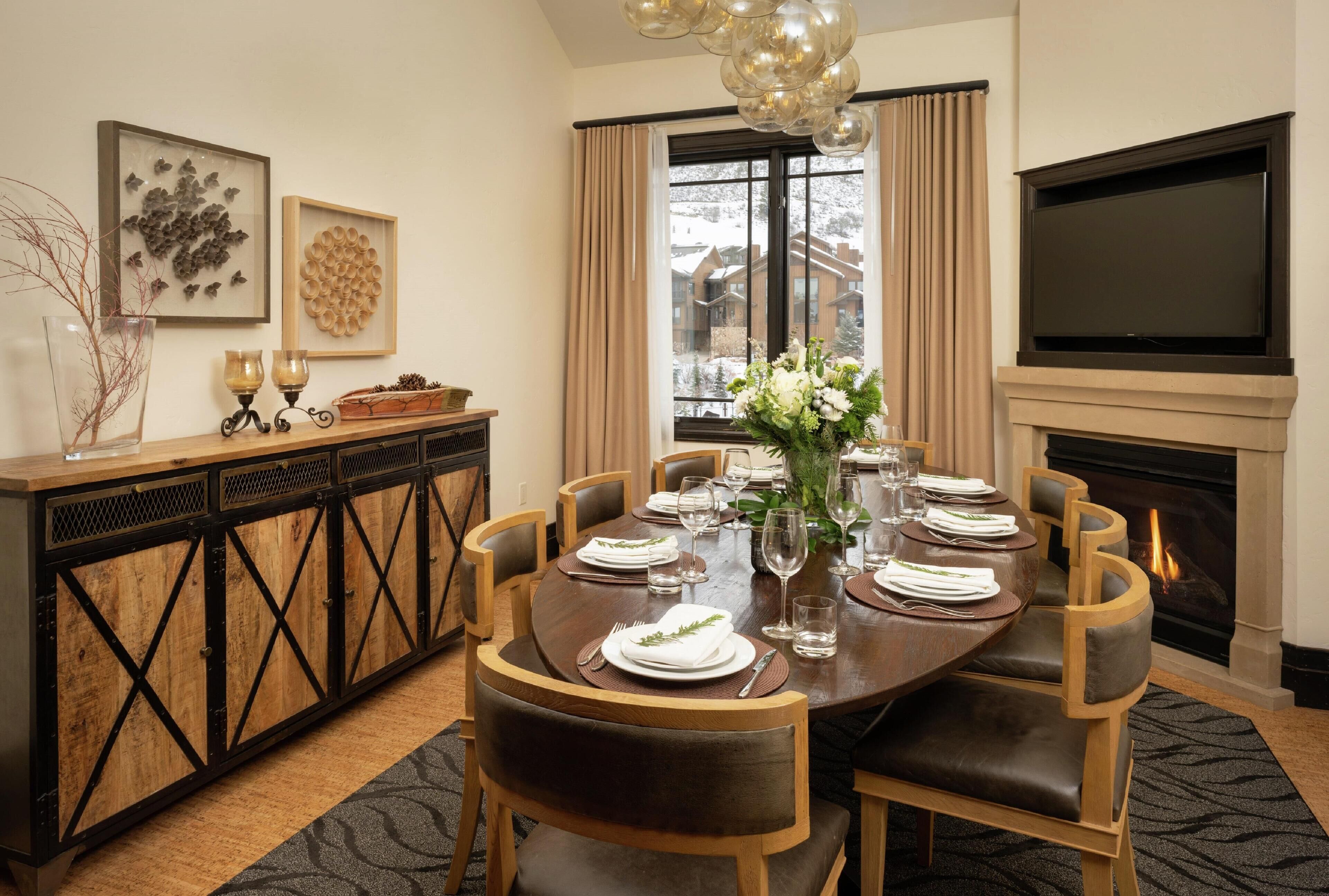 A dining room with a round table set for six, flanked by wooden chairs with leather cushions. A fireplace is visible to the right, and a large flat-screen TV is mounted on the wall above it.