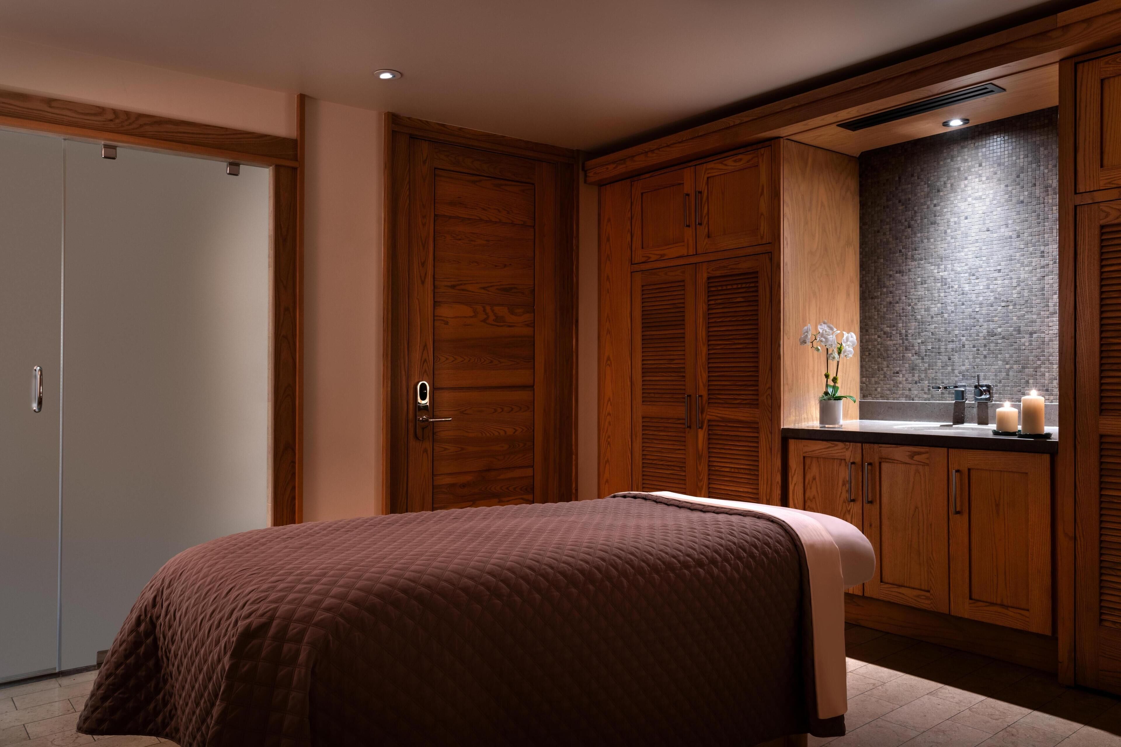 A bedroom with a large bed, wooden wardrobe, and a kitchenette area with a brick backsplash.