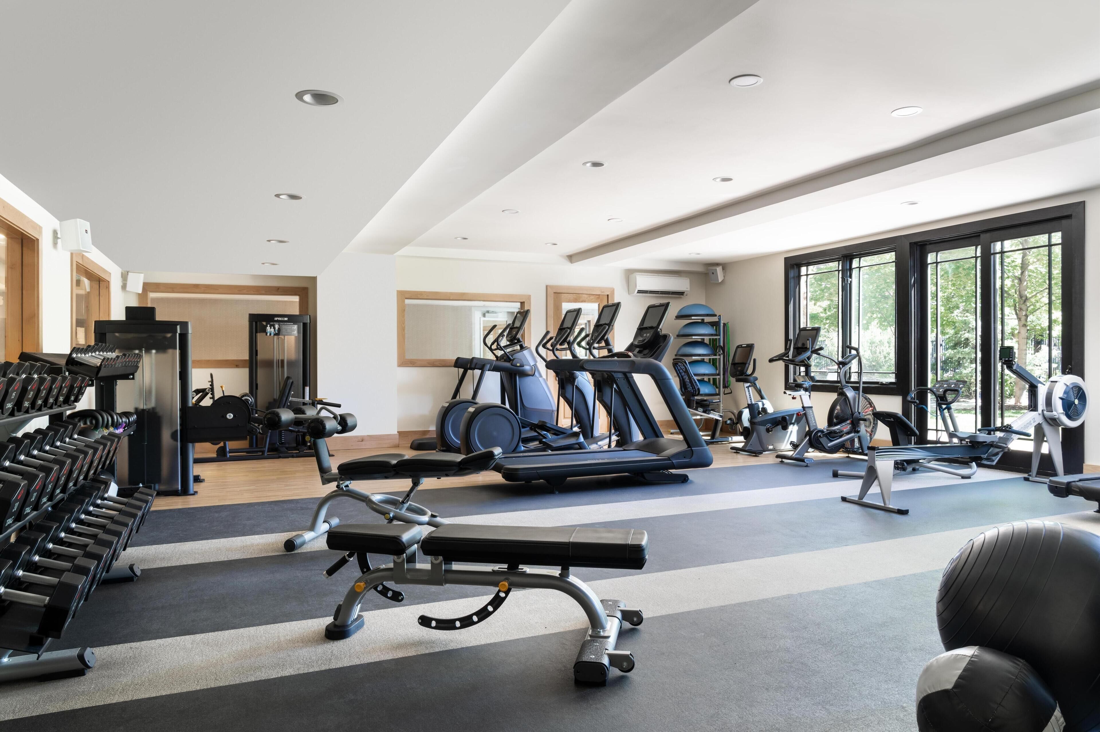 A modern gym with various exercise equipment, large windows, and a clean, spacious layout.
