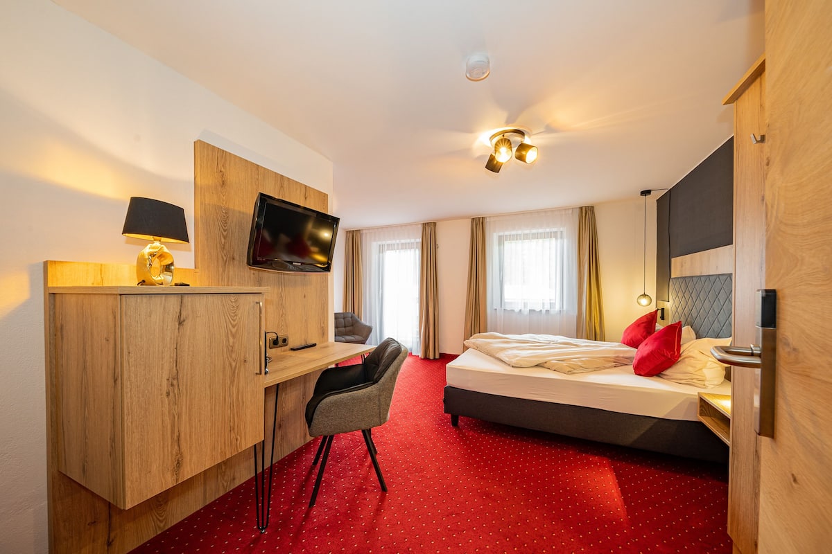 Une chambre d’hôtel avec un bureau en bois, une télévision, un lit avec des oreillers rouges et une moquette rouge.