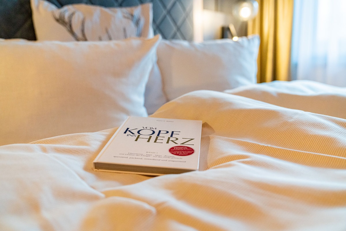 Un livre intitulé « KOPE HERZ » est posé sur un lit recouvert d’une literie blanche et agrémenté d’oreillers.