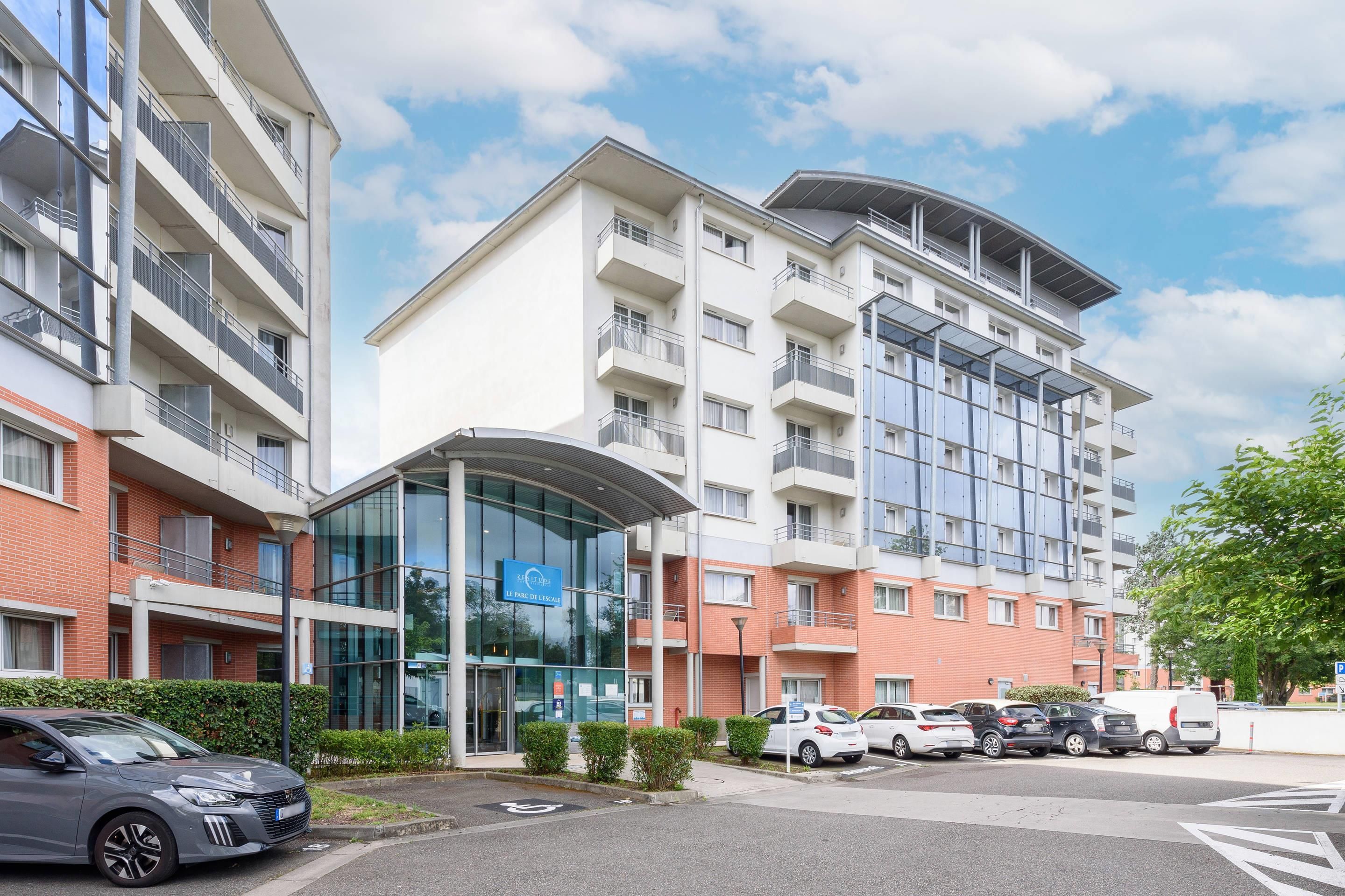 A modern multi-story building with a glass entrance, parking area, and several parked cars.