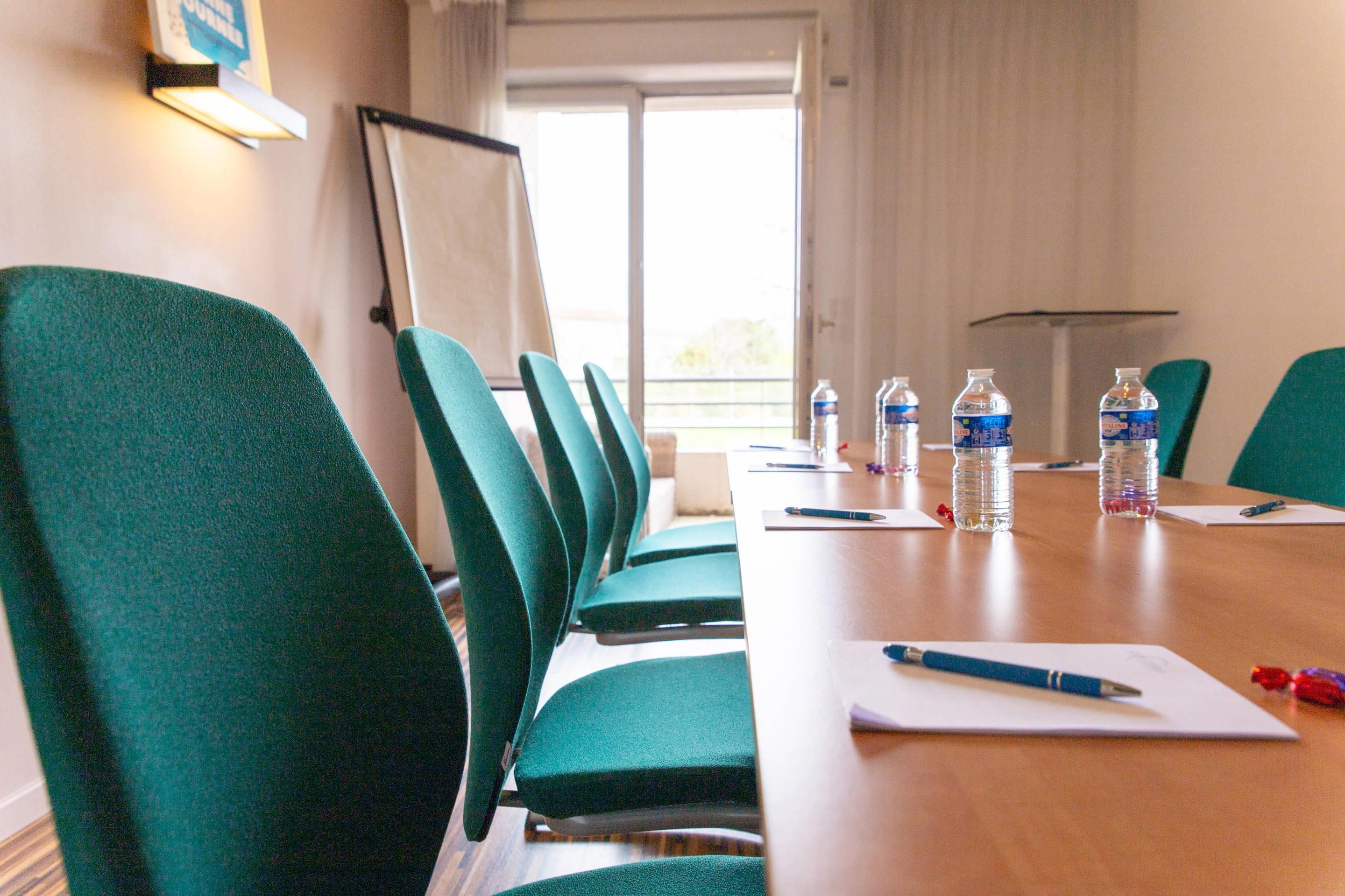 Ein Konferenzraum mit grünen Stühlen, einem langen Tisch, Wasserflaschen und einer Whiteboard-Tafel.