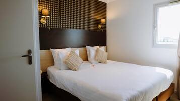 Una cama impecablemente vestida con sábanas blancas y dos almohadones decorativos, cabecera oscura con un diseño geométrico y dos lámparas fijadas a la pared.