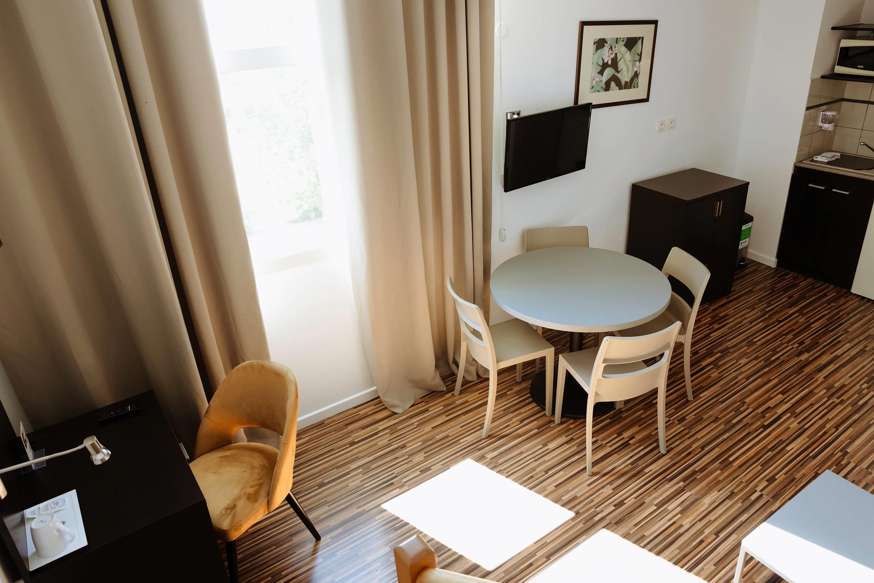 Une chambre d’hôtel avec une table de salle à manger ronde, deux chaises, un petit coin cuisine, un téléviseur fixé au mur et une fenêtre avec des rideaux.