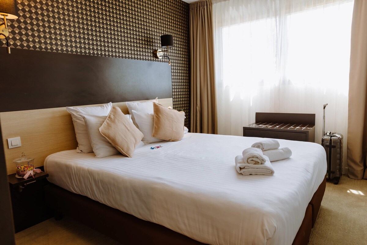 A neatly made bed with white linens and several pillows, a bedside table with a lamp, a suitcase, and a wall with a geometric pattern.