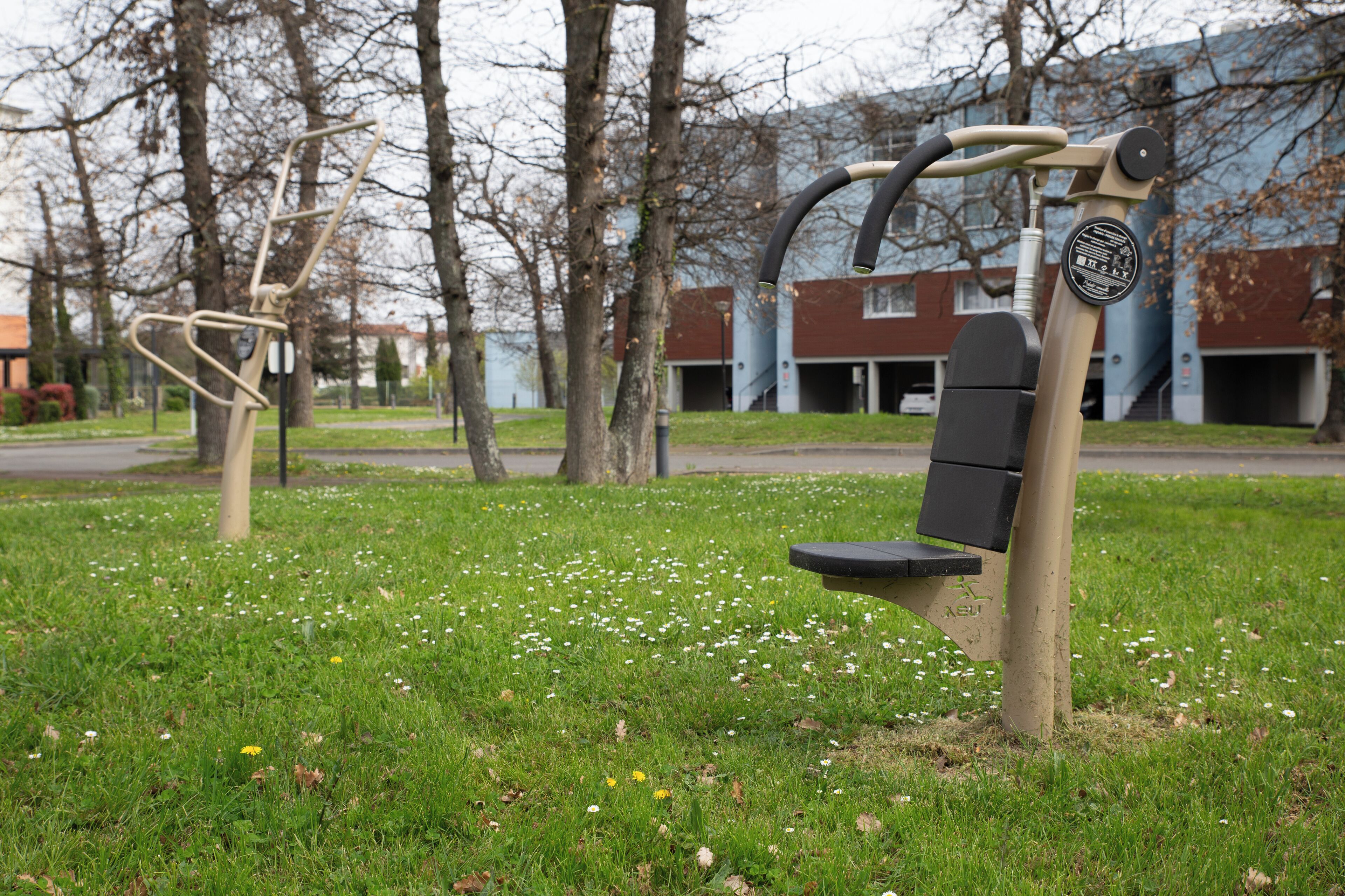 Outdoor exercise equipment in a grassy area with trees and buildings in the background.