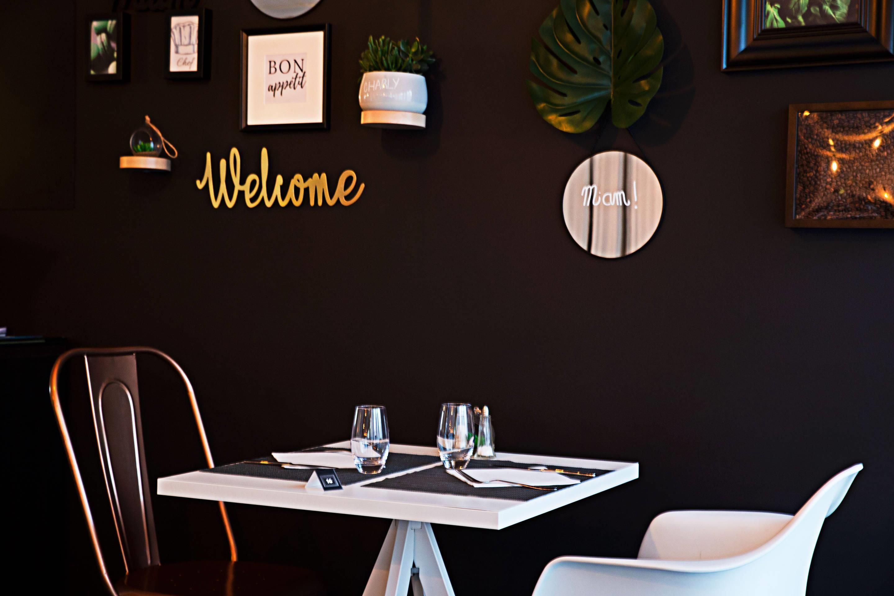 Ein Essbereich mit einem Tisch für zwei Personen, einem Stuhl und einer Wand mit Dekoration, darunter ein Schild mit der Aufschrift 'Welcome' und gerahmte Bilder.