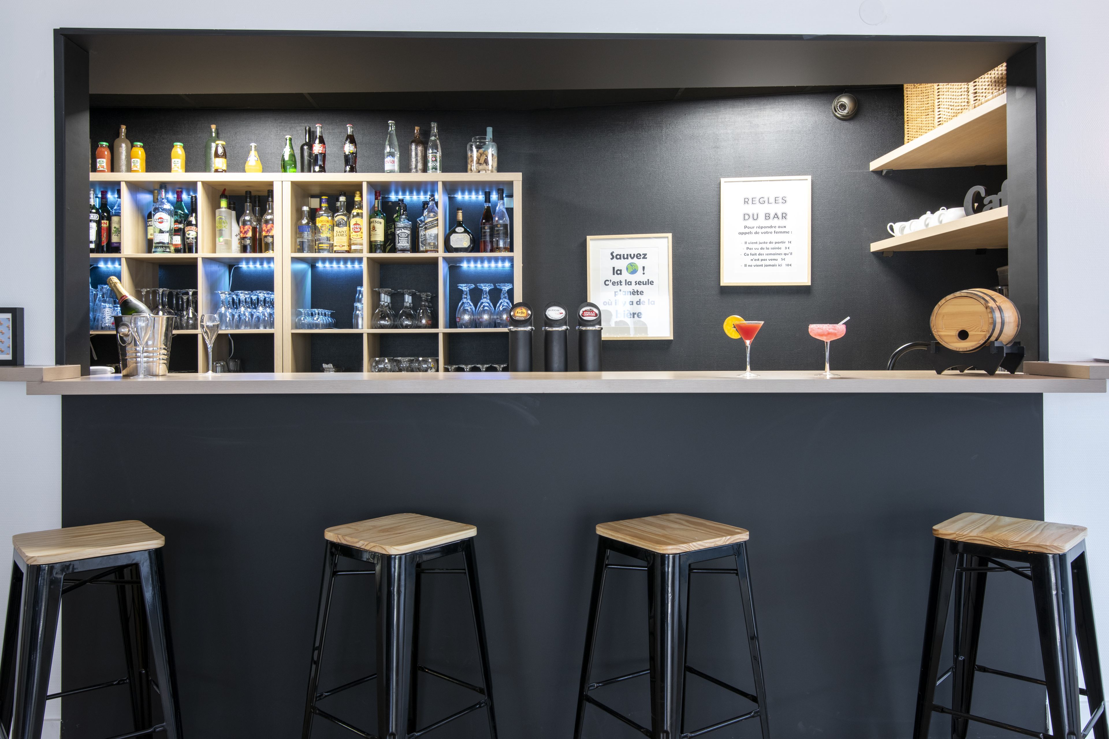 Zona de bar con pared oscura, estantes llenos de botellas de licor y dos taburetes.