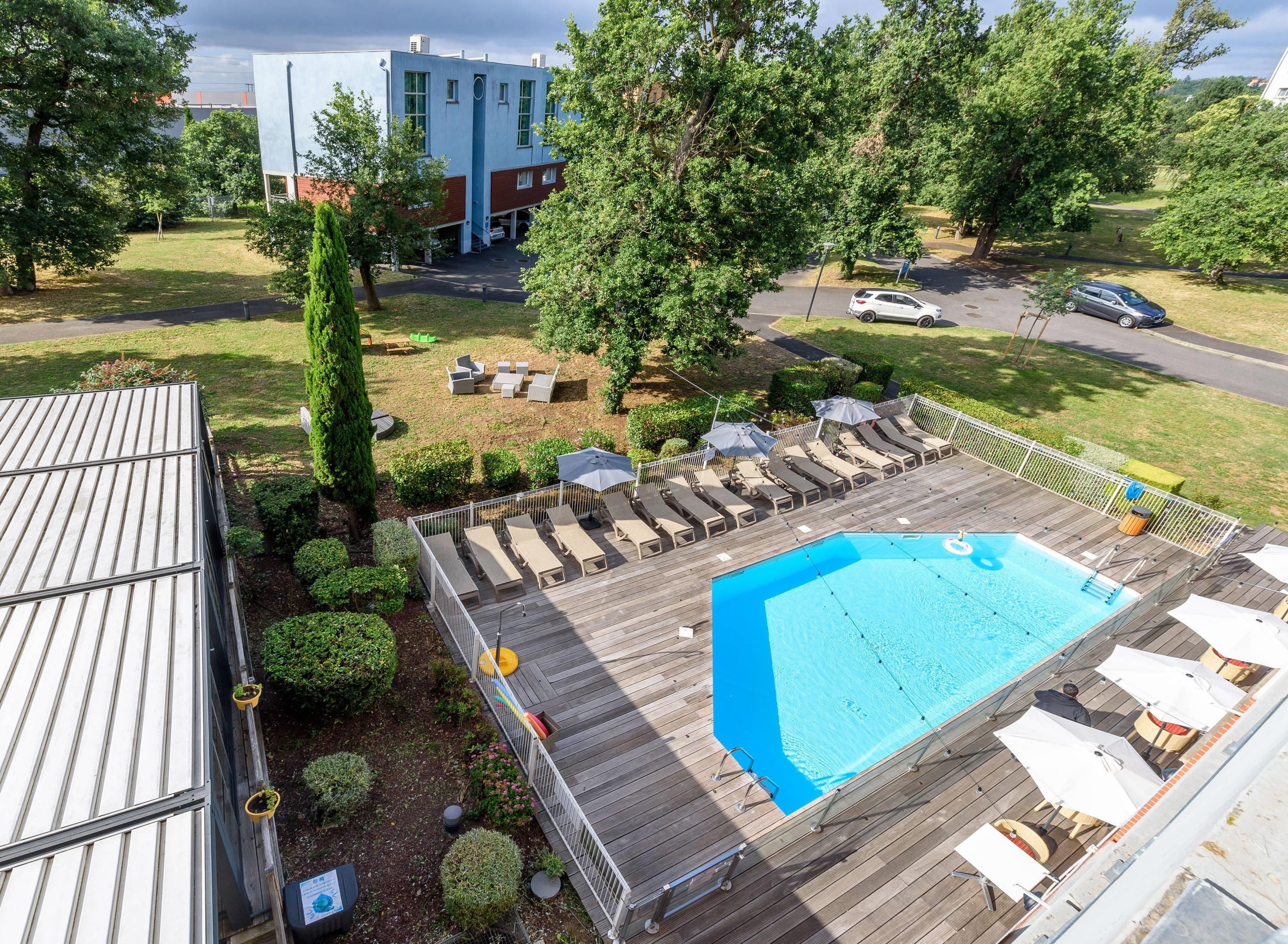 Une piscine sur le toit, dotée de chaises longues, d’une terrasse en bois et offrant une vue sur un bâtiment en arrière-plan.