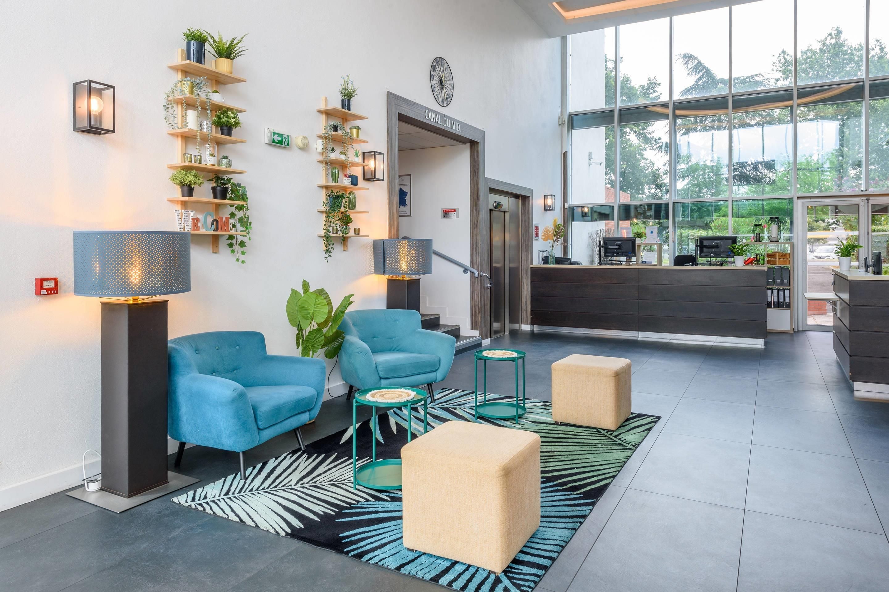 A modern lobby with a reception desk, blue armchairs, a large window, and a plant wall.