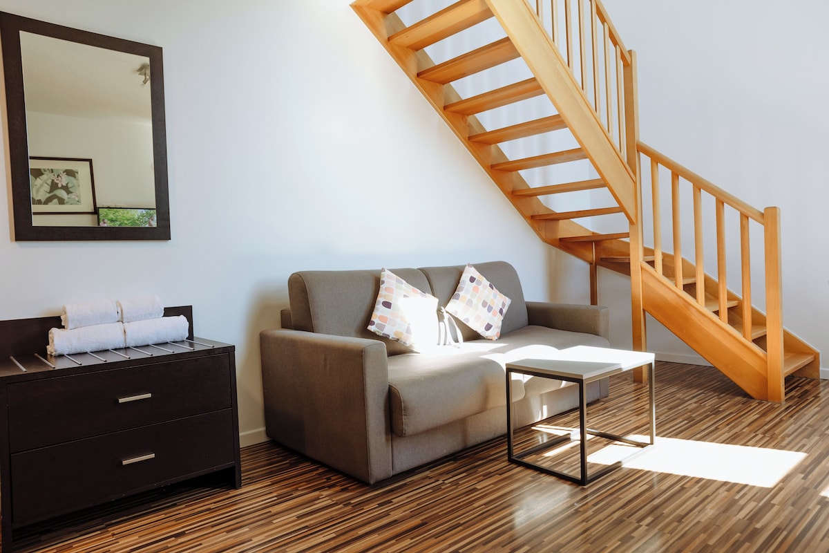 A modern living room with a wooden staircase, a sofa with cushions, a side table, and a dark wooden dresser.