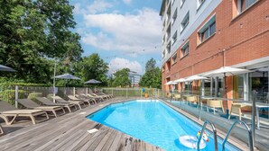Une piscine entourée d’un pont en bois et d’un bâtiment en briques, avec des chaises longues et des parasols à proximité.