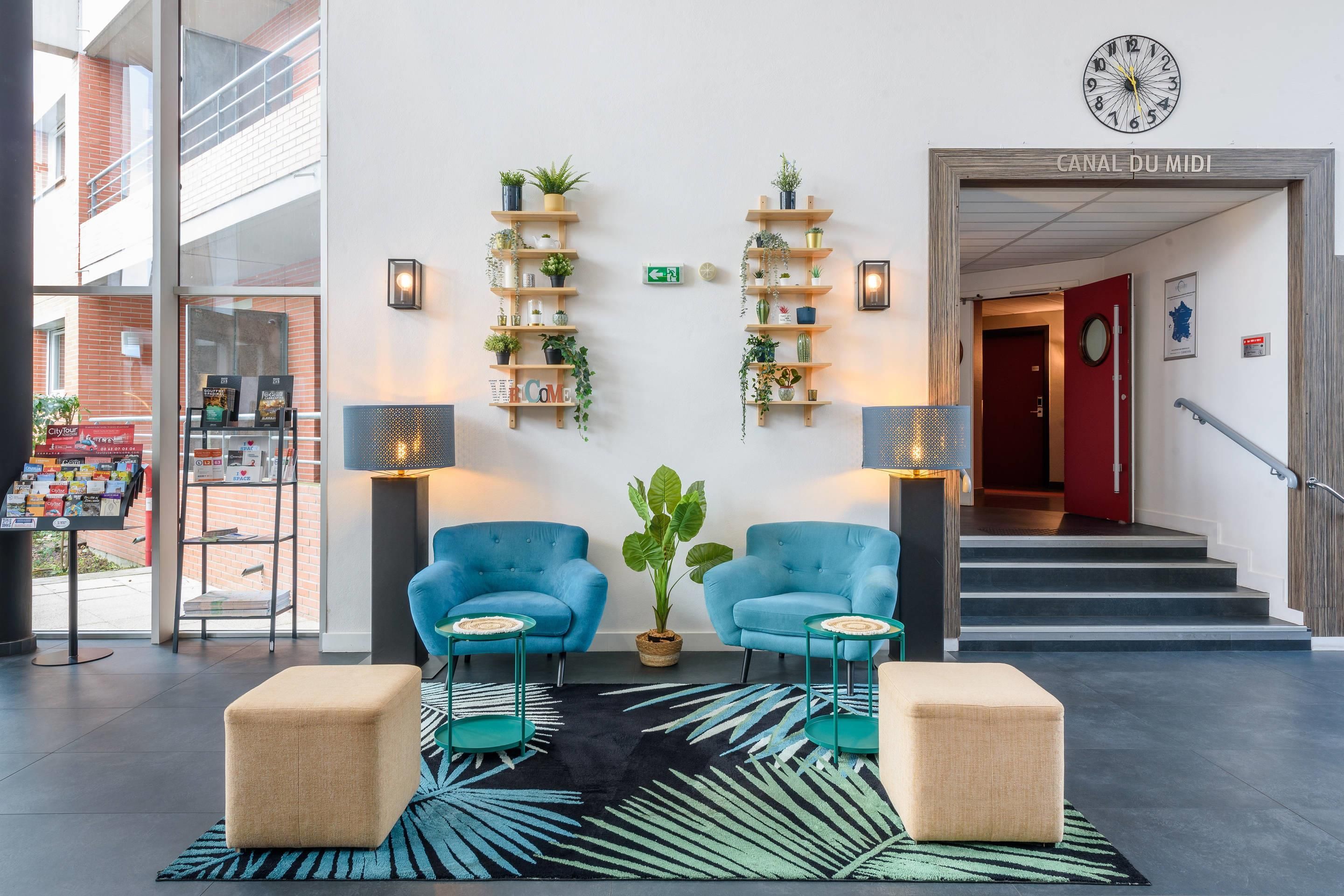 Un vestíbulo con dos sillones azules, un tapete con estampado negro y verde, y estanterías de madera con plantas.