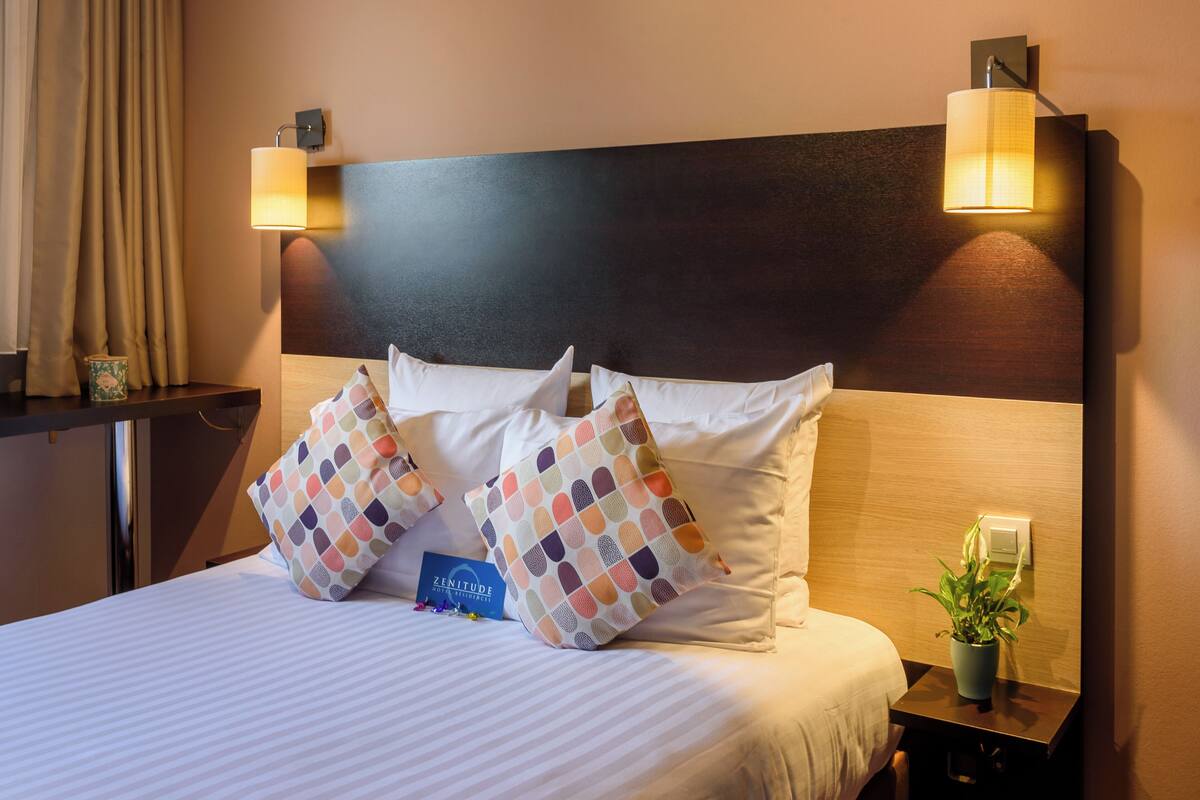 Une chambre d’hôtel avec un lit, deux oreillers, une table de chevet avec une lampe, une plante et un panneau posé sur la table de chevet.