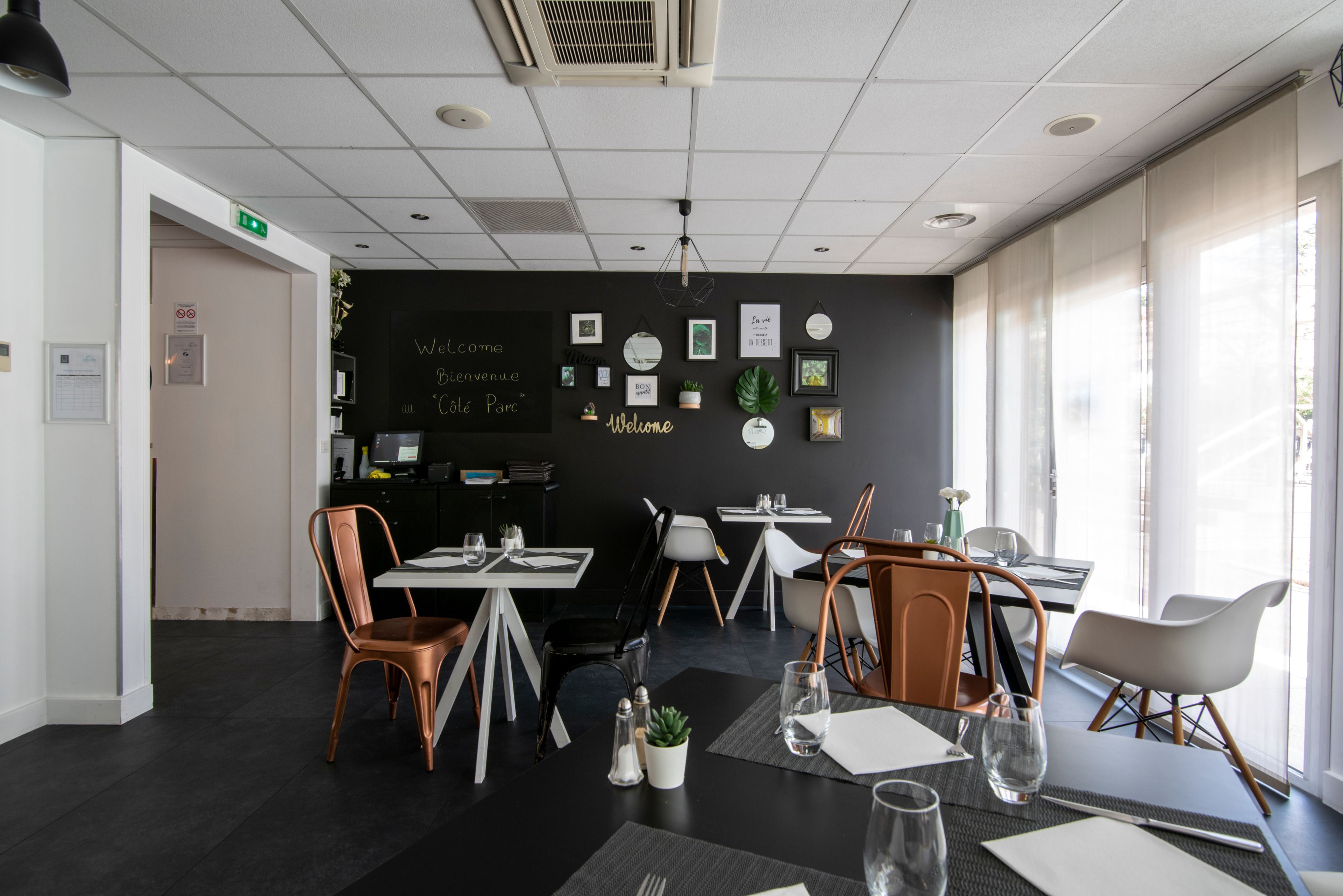Un comedor moderno con mesas y sillas, una pared de pizarra con los mensajes 'Welcome' y 'Bienvenue', y vistas al exterior a través de grandes ventanales.